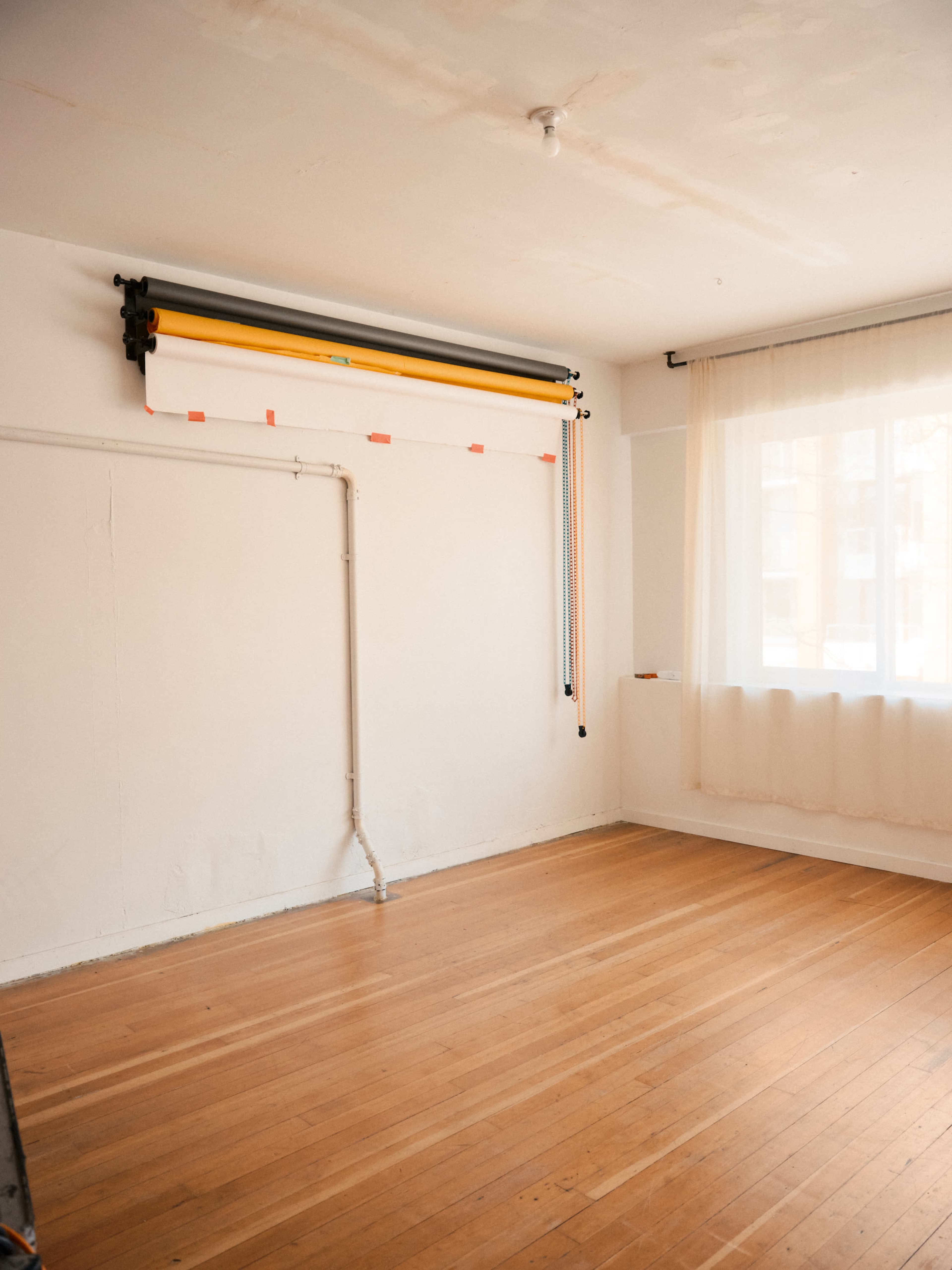 An empty room features wooden flooring, a large window with sheer curtains, and a wall-mounted backdrop system.