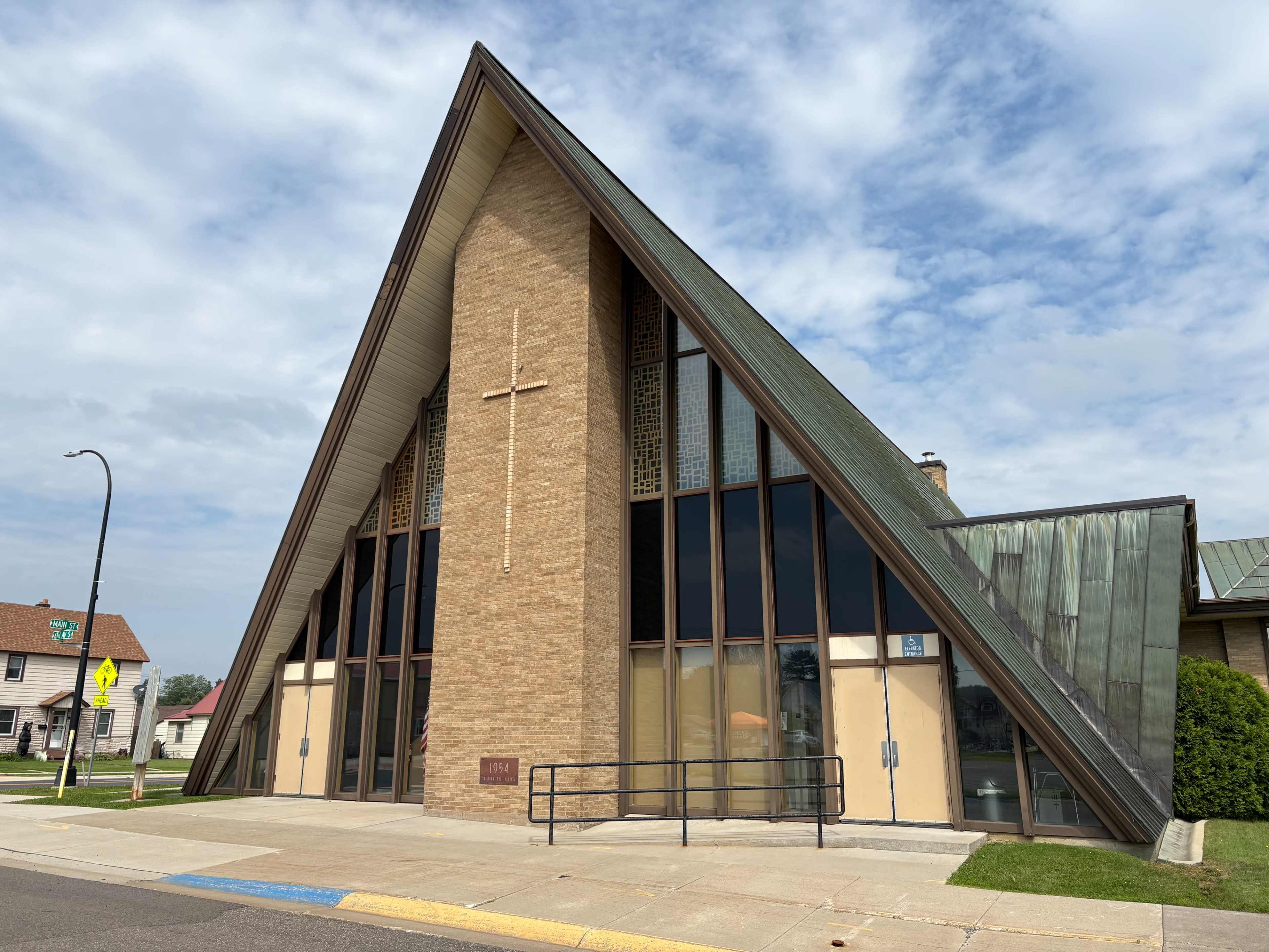 The image shows a modern church building with a distinctive triangular roof and large windows, situated on a street corner.