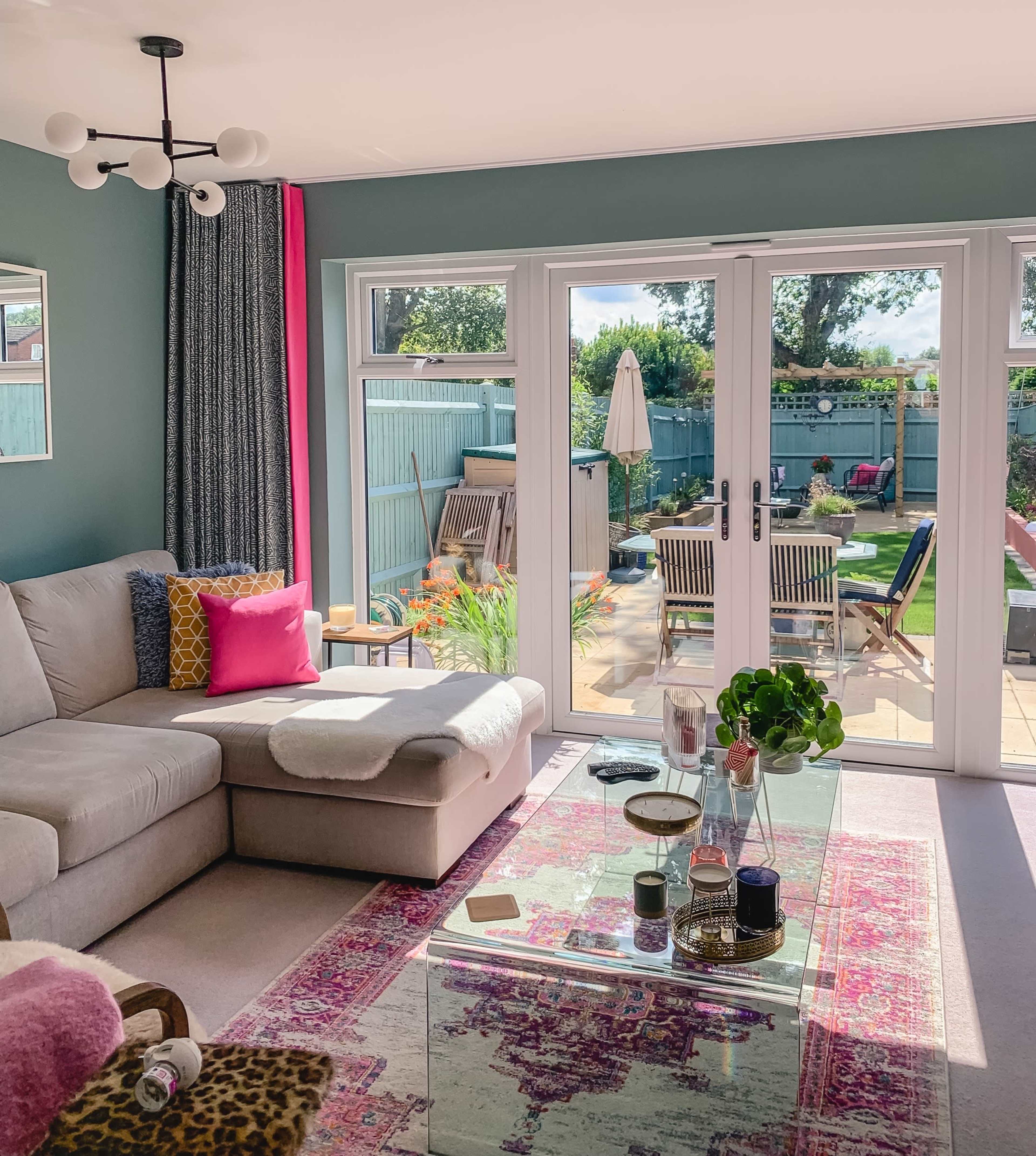 A bright living room with a large window overlooking a garden, featuring a beige sectional sofa, a glass coffee table, and vibrant decor.