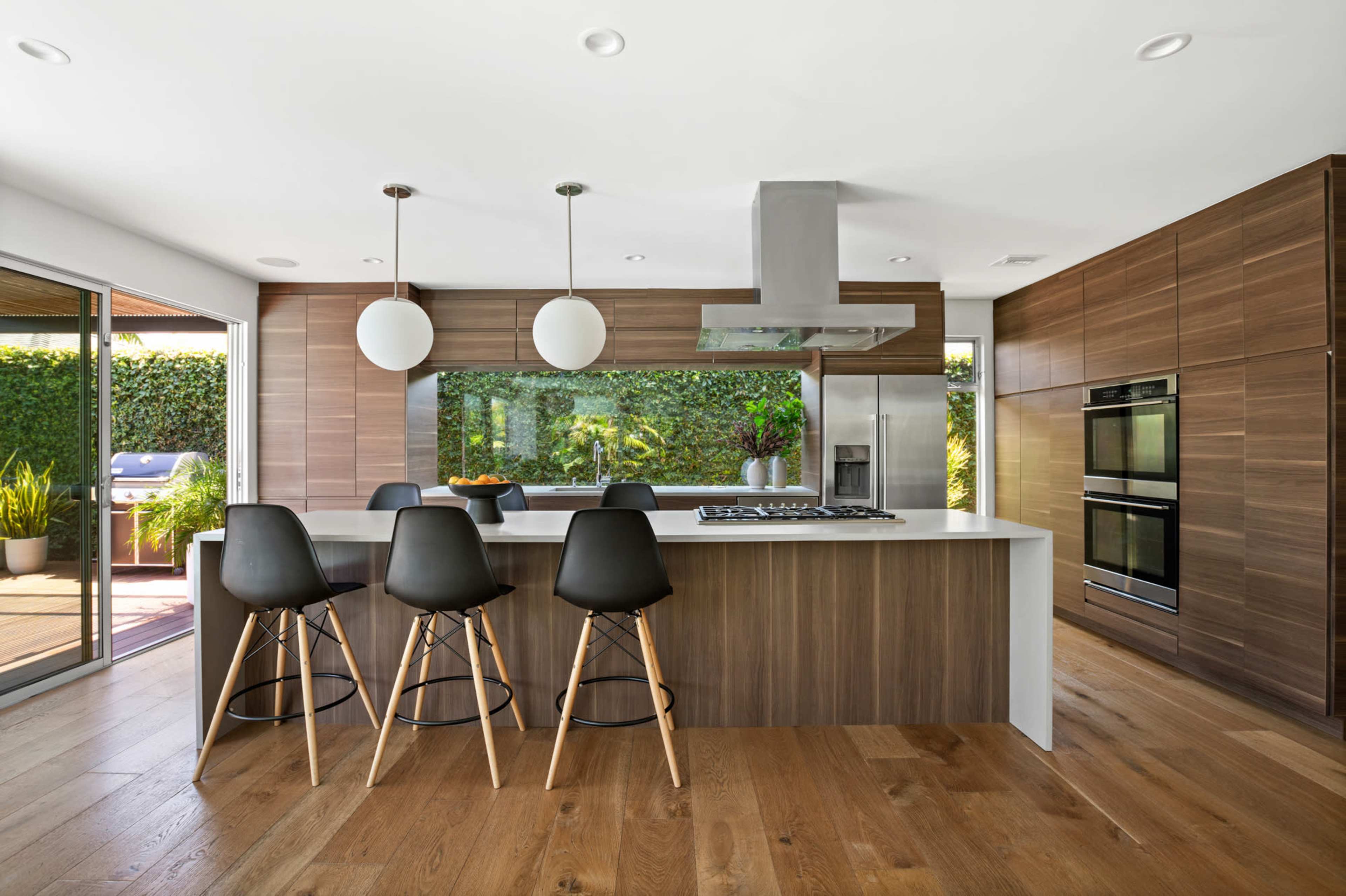 A modern kitchen features a large island with black stools, wooden cabinetry, and stainless steel appliances, surrounded by greenery visible through glass doors.