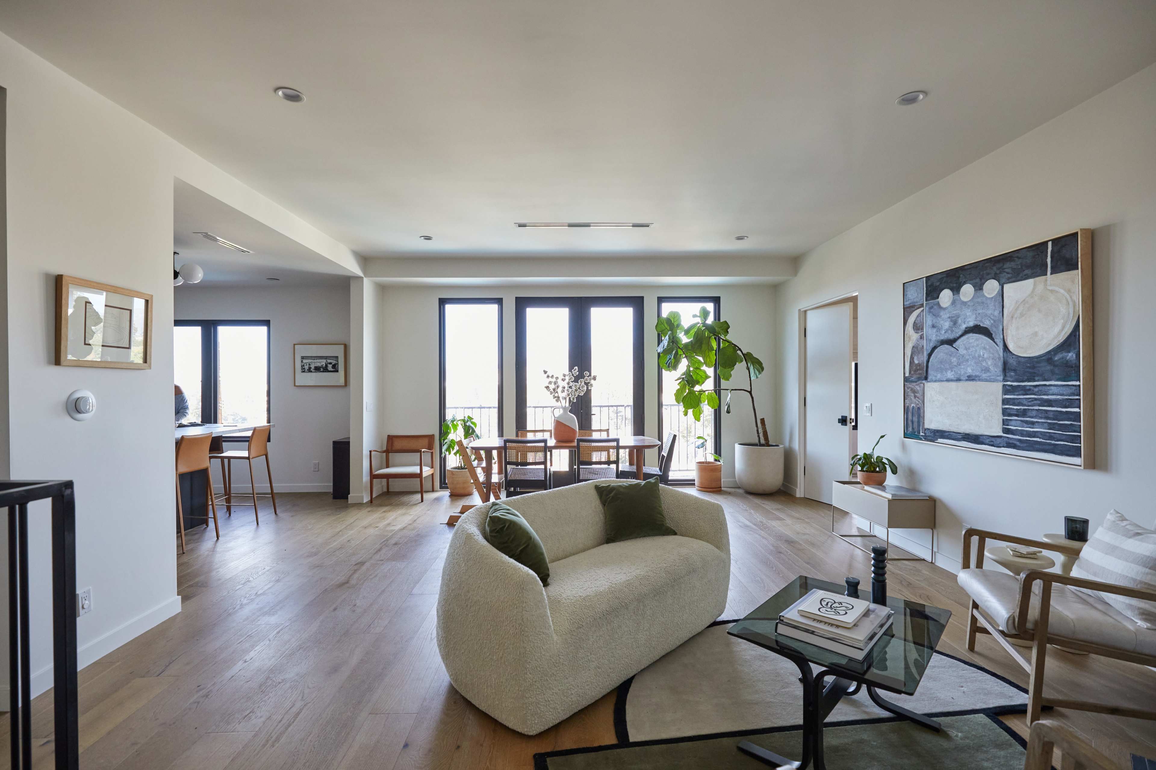 The image shows a modern living room with light wood flooring, large windows, and a mix of stylish furniture including a curved sofa and a glass coffee table.