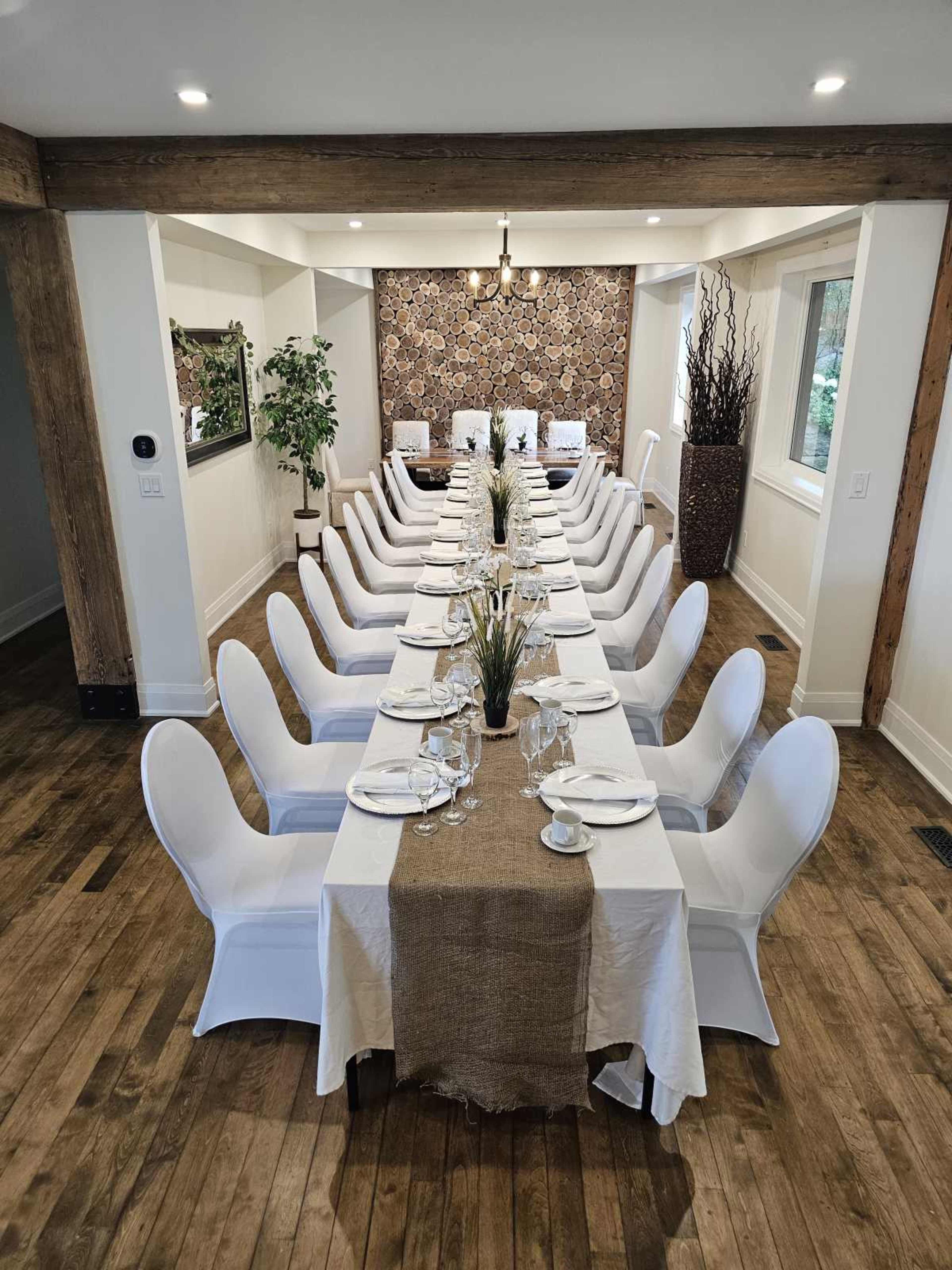 A long dining table is elegantly set with white chairs and decorative centerpieces in a well-lit room featuring wooden beams and a patterned accent wall.