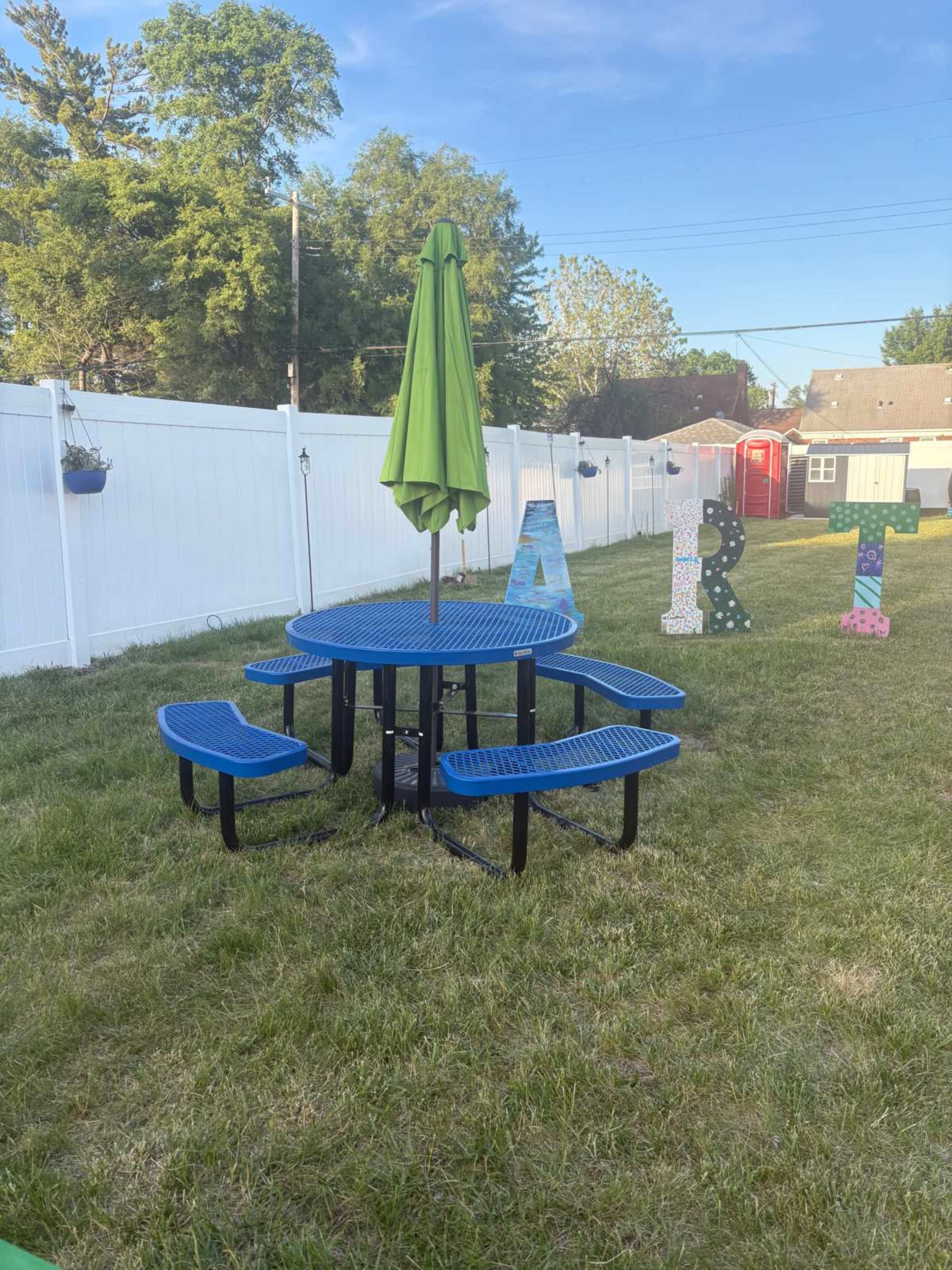 A blue picnic table with attached benches and a green umbrella is situated in a grassy backyard, surrounded by a white fence and colorful letter decorations.