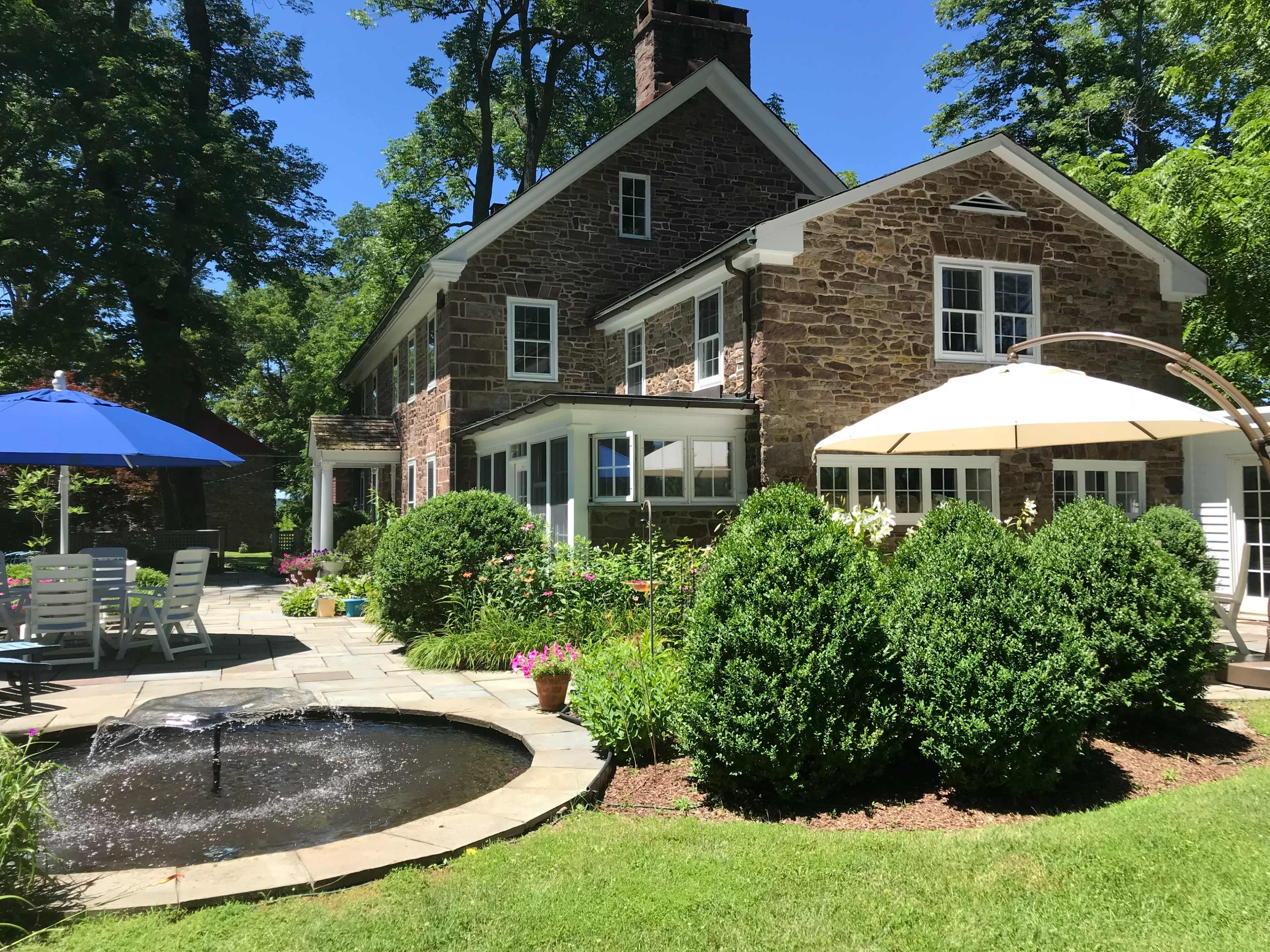 A stone house is surrounded by well-maintained gardens and a small pond with a fountain, featuring patio furniture and umbrellas for outdoor seating.