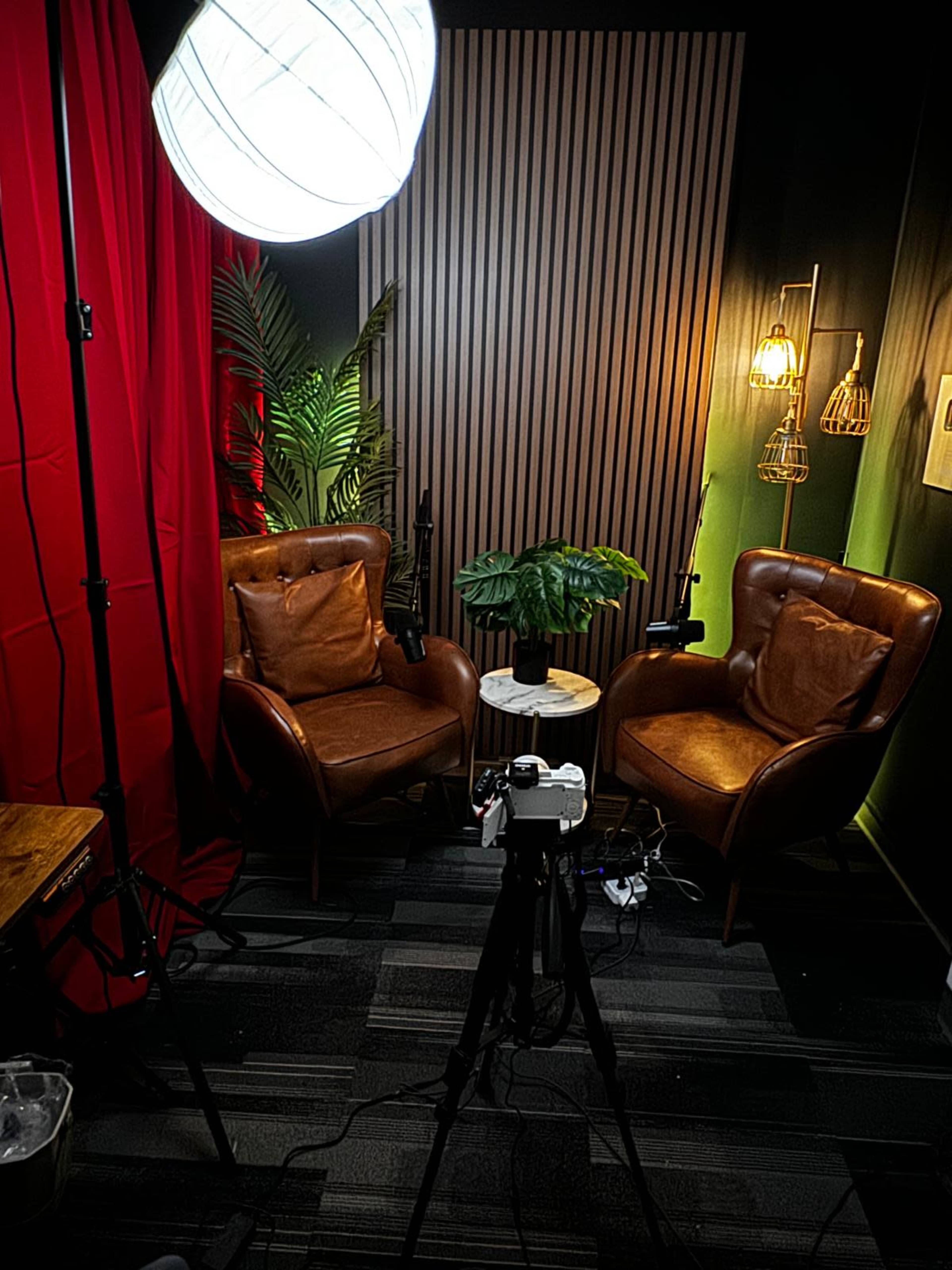 The image shows a cozy setup featuring two leather armchairs, a small table, and a camera on a tripod, all arranged in a room with red curtains and a well-lit background.
