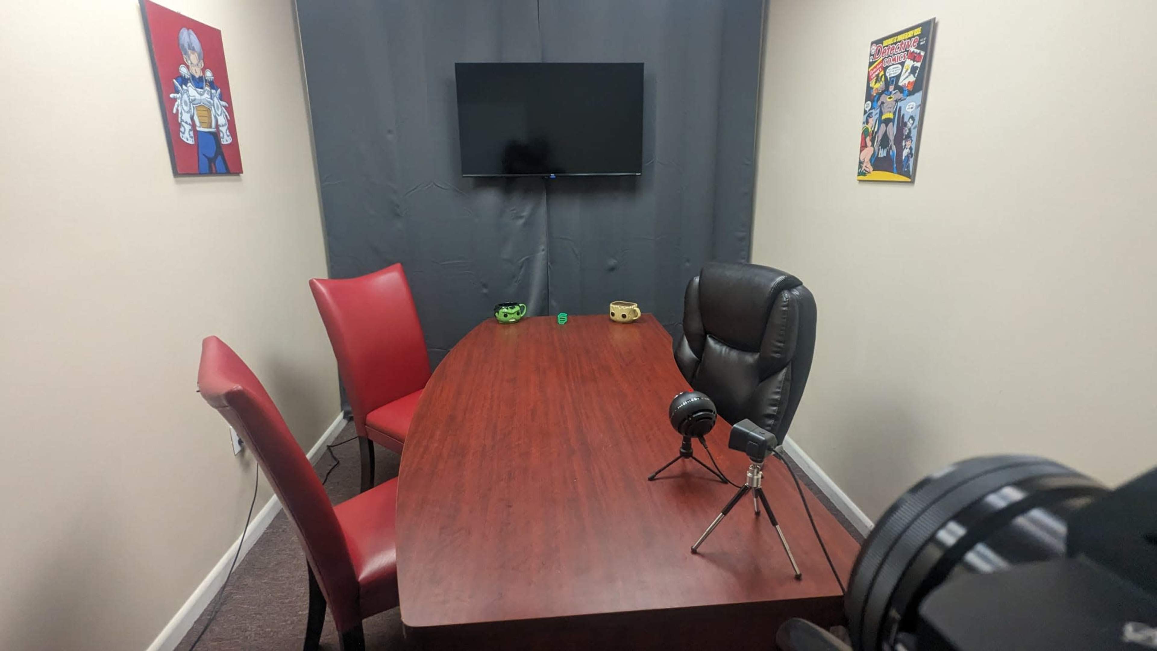 The image shows a small, enclosed meeting room featuring a wooden table, two chairs, a wall-mounted television, and comic book posters on the walls.