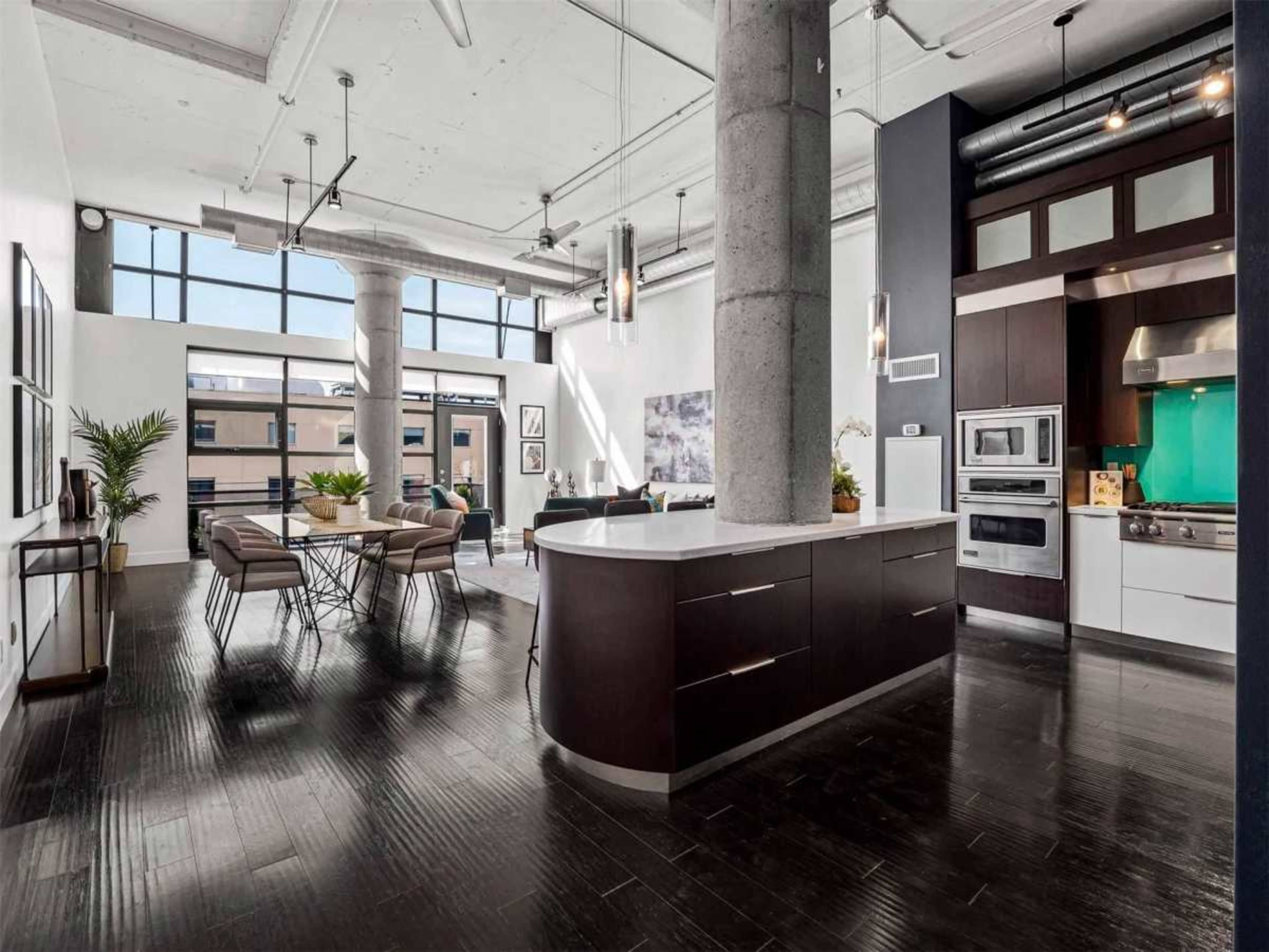 The image shows a modern open-concept living space with a kitchen, dining area, and seating area, featuring large windows and industrial-style elements.