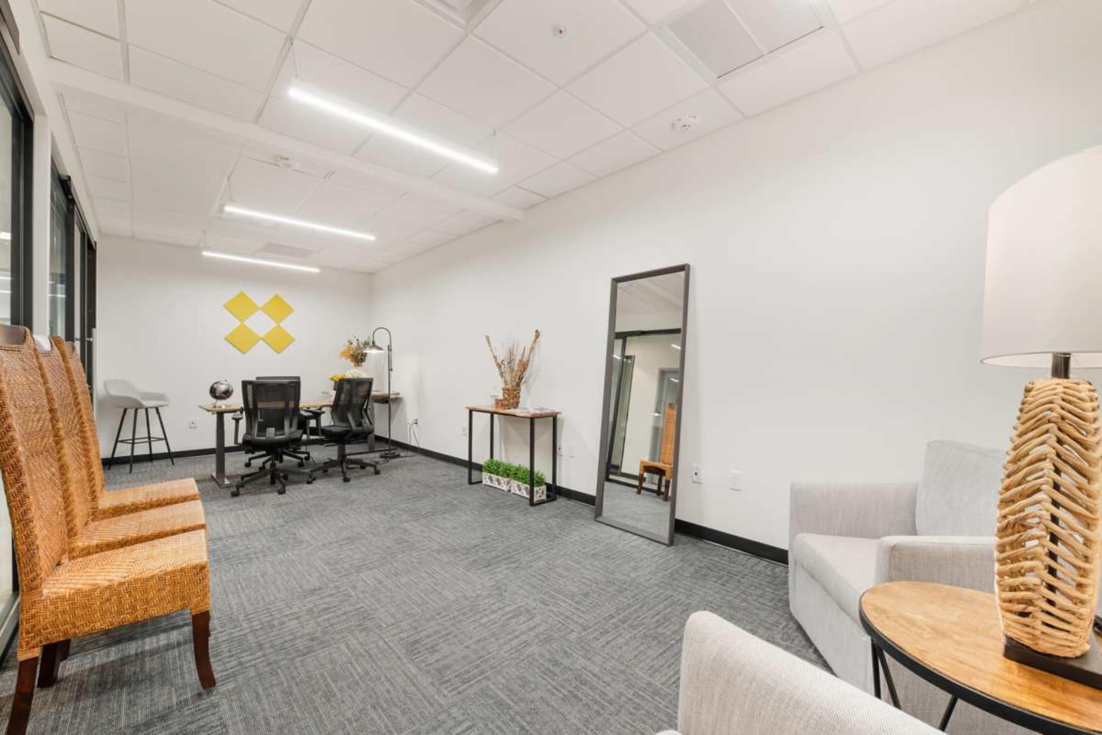The image shows a modern office space with two seating areas, a desk with office chairs, a large mirror, and decorative elements.