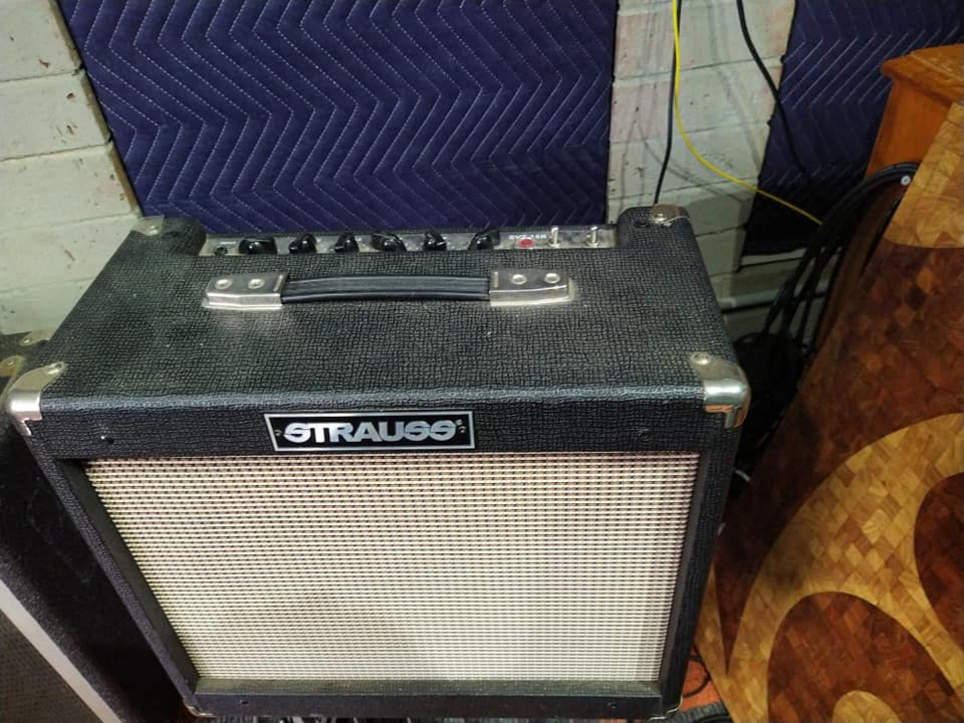 A black guitar amplifier with control knobs on the top and a metal handle, placed on a textured surface in a room.