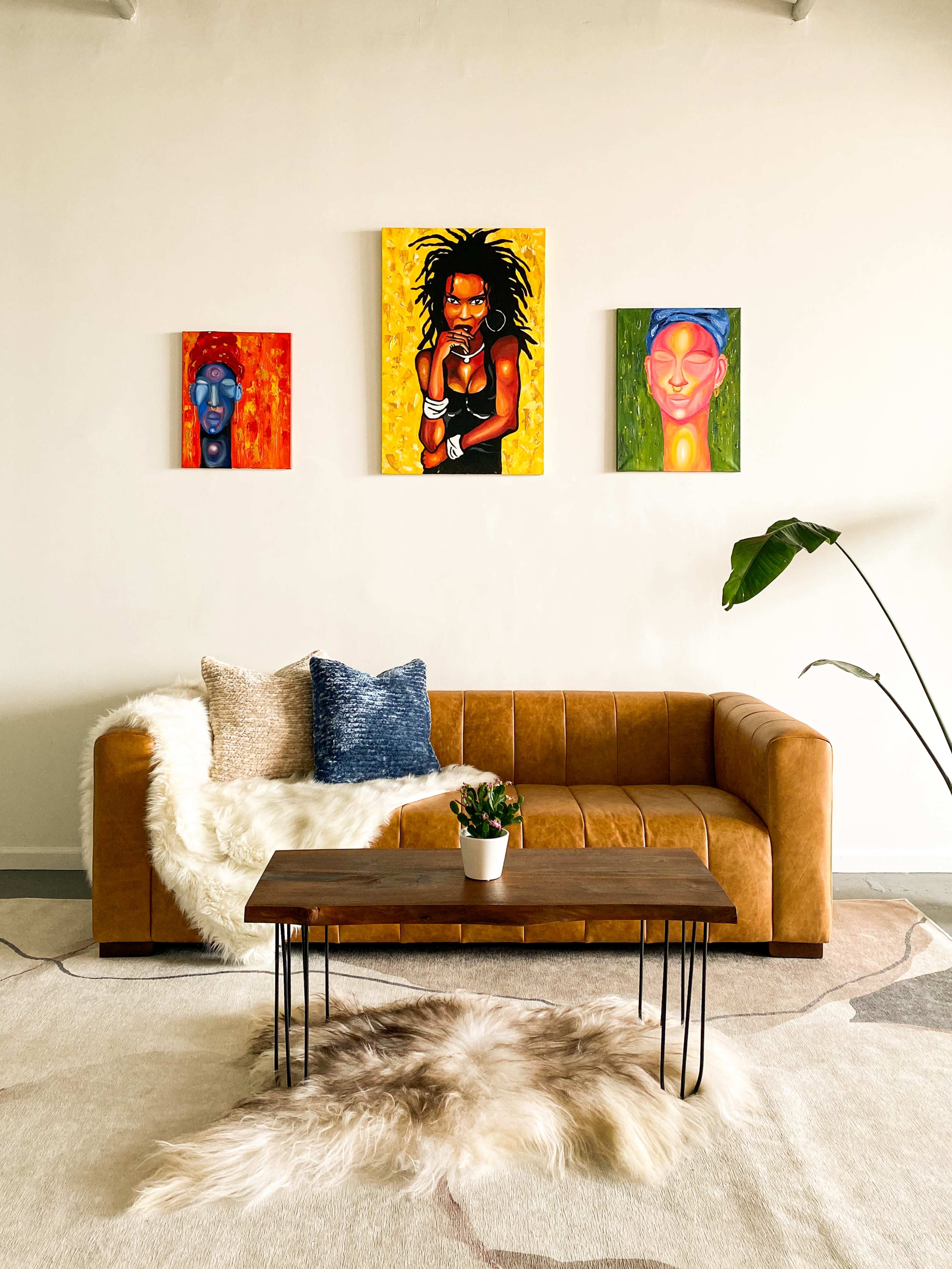 A cozy living room features a brown leather sofa, a wooden coffee table, and three colorful paintings on the wall, with a pile of faux fur and a potted plant nearby.