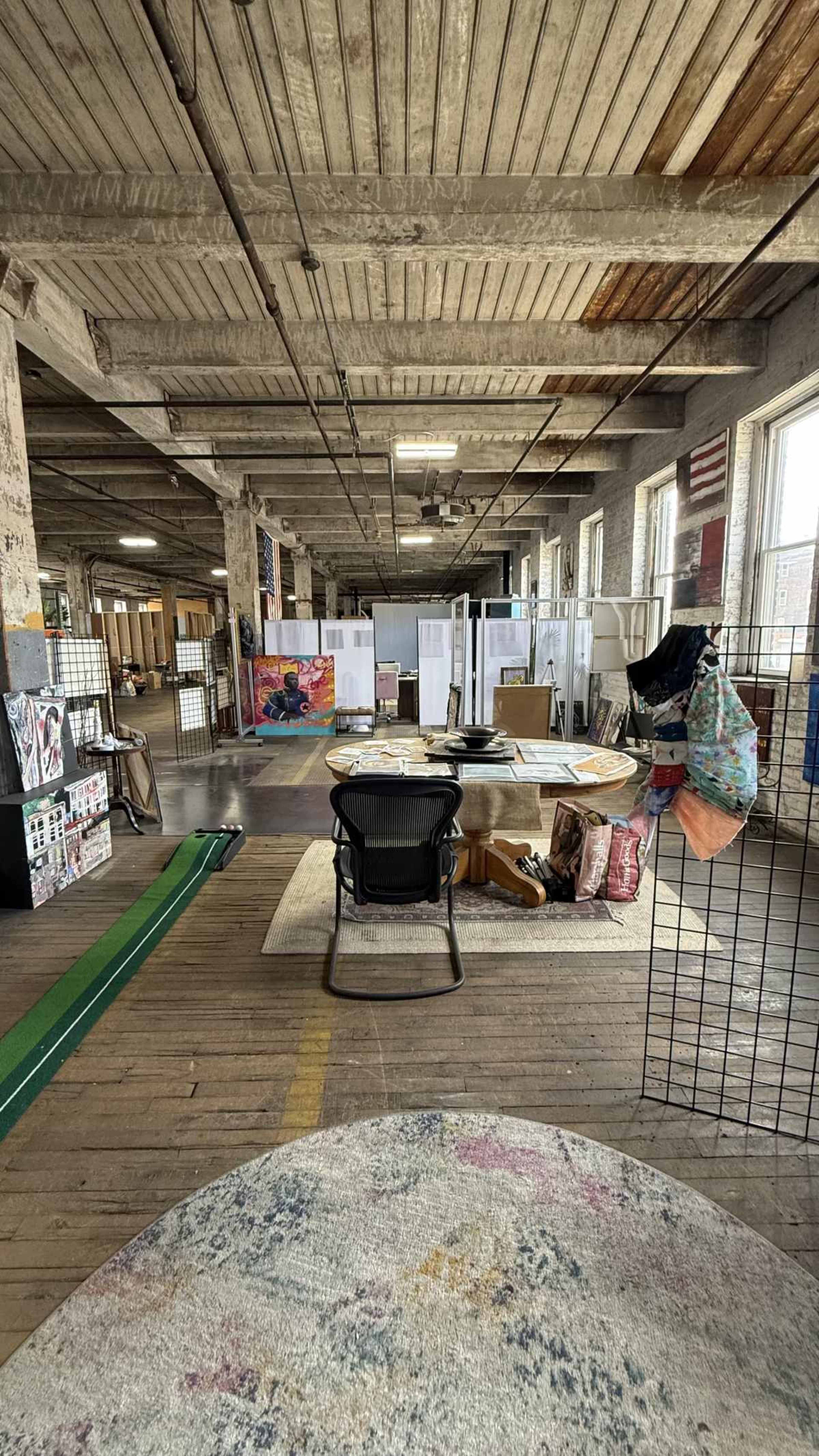 The image shows a spacious, industrial-style workspace featuring wooden floors, exposed beams, and various art supplies and furniture scattered around.