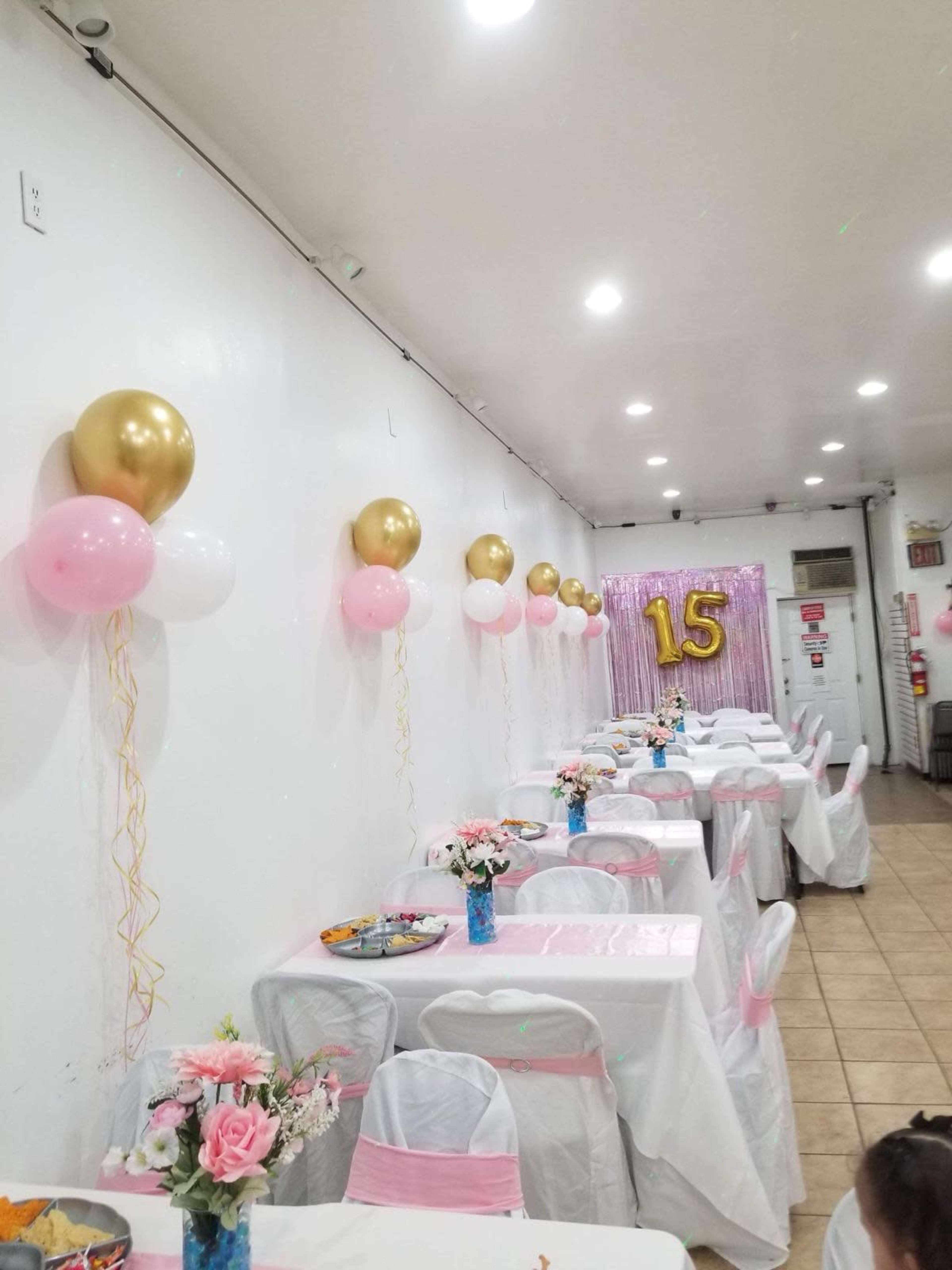 A party setting with white tables covered in tablecloths, decorated with pink and gold balloons along the walls, and floral centerpieces.
