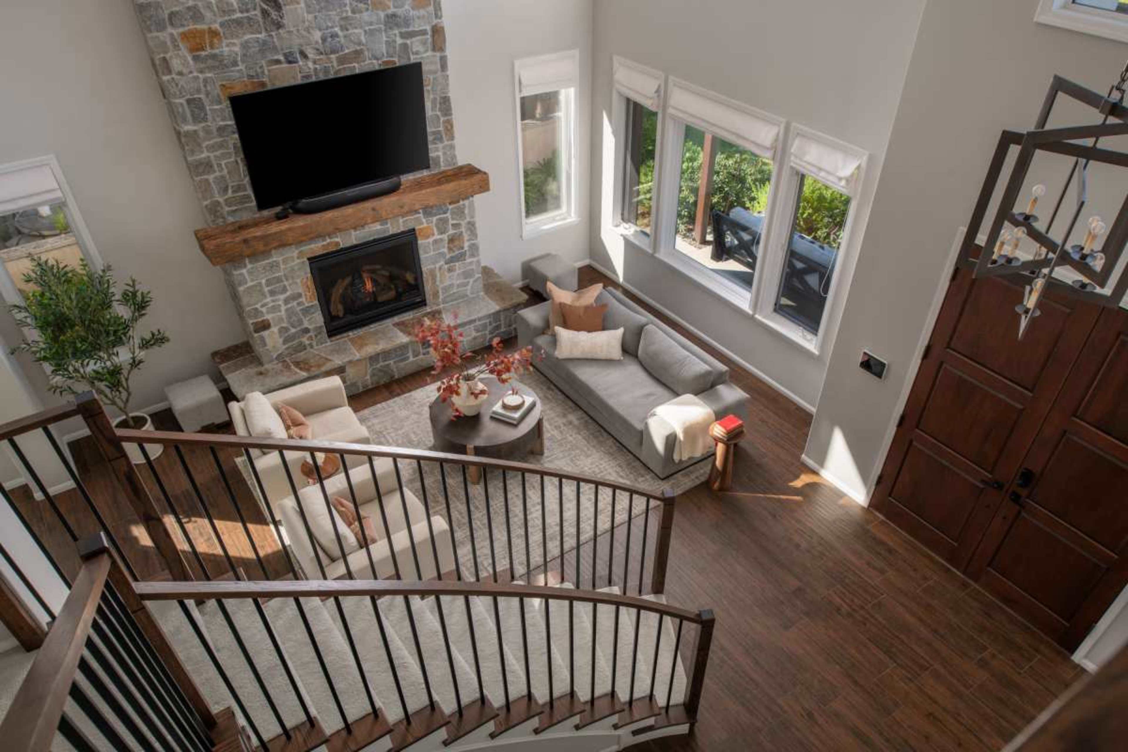 The image shows a spacious living room featuring a stone fireplace, a large sectional sofa, and a decorative coffee table, all arranged around a central area rug with large windows letting in natural light.