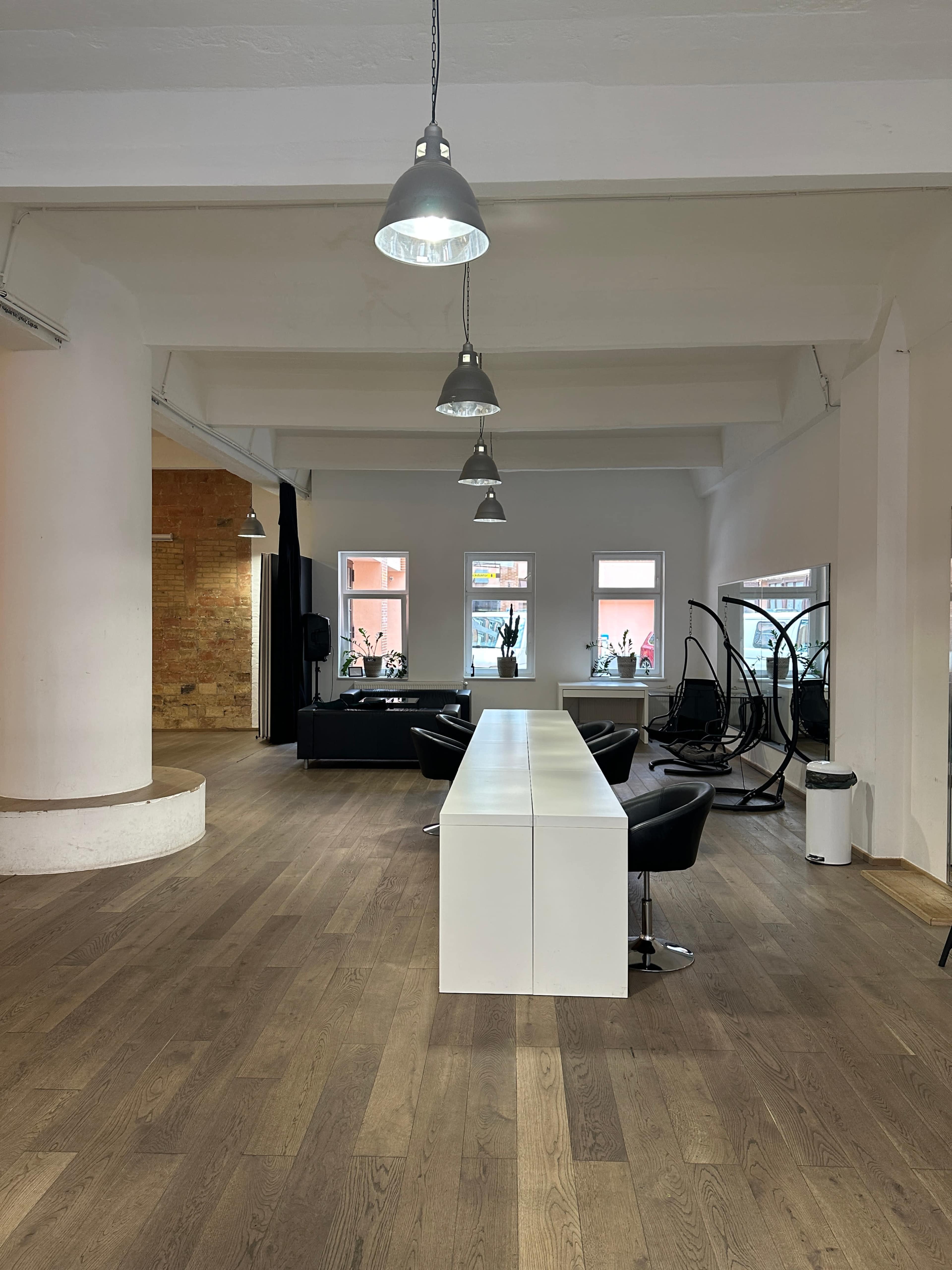 The image shows a spacious, modern interior featuring a long white table, black chairs, large windows, and wooden flooring.