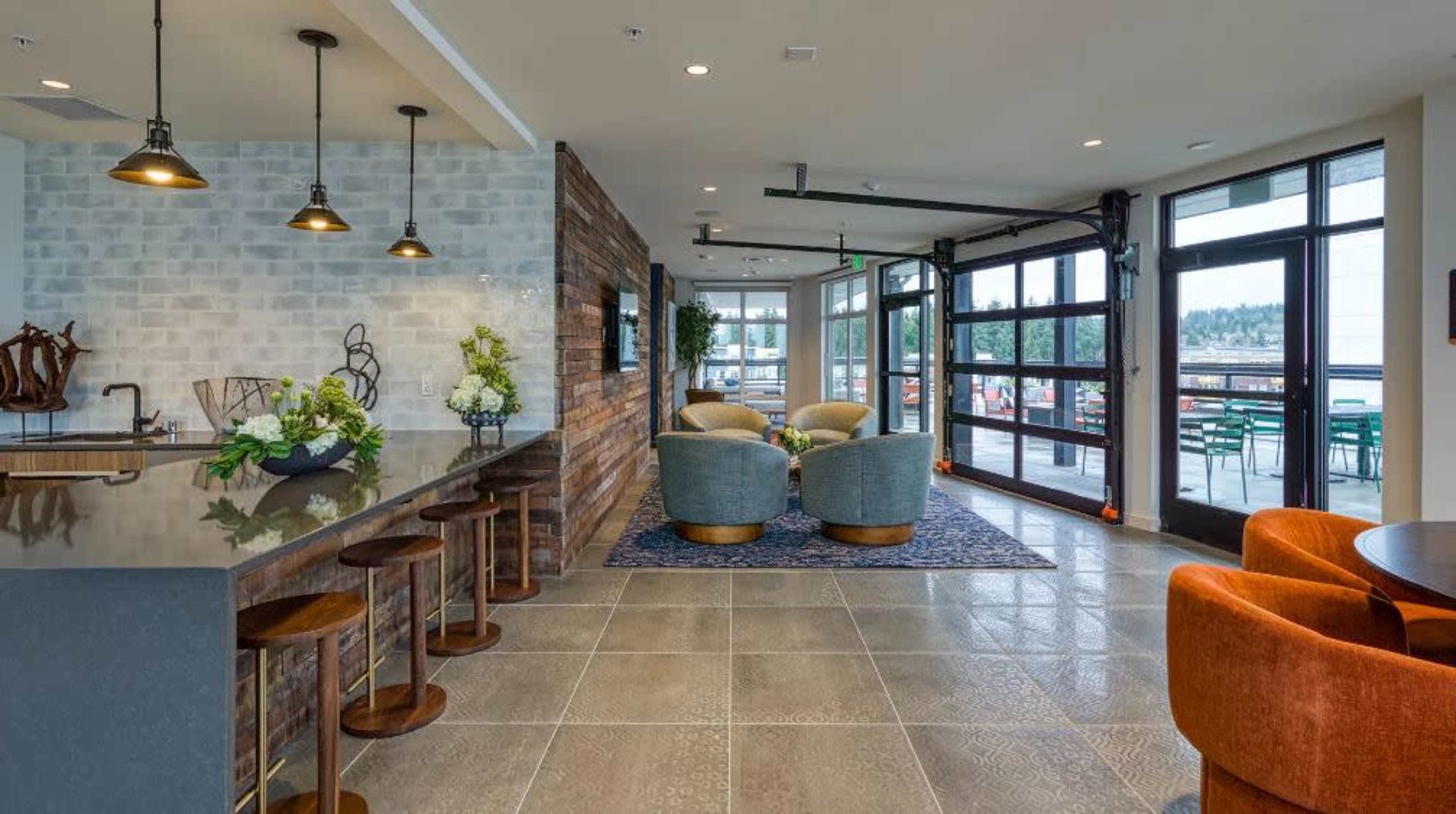 A modern interior space featuring a bar area with stools, a seating area with two chairs, and large windows overlooking an outdoor patio.