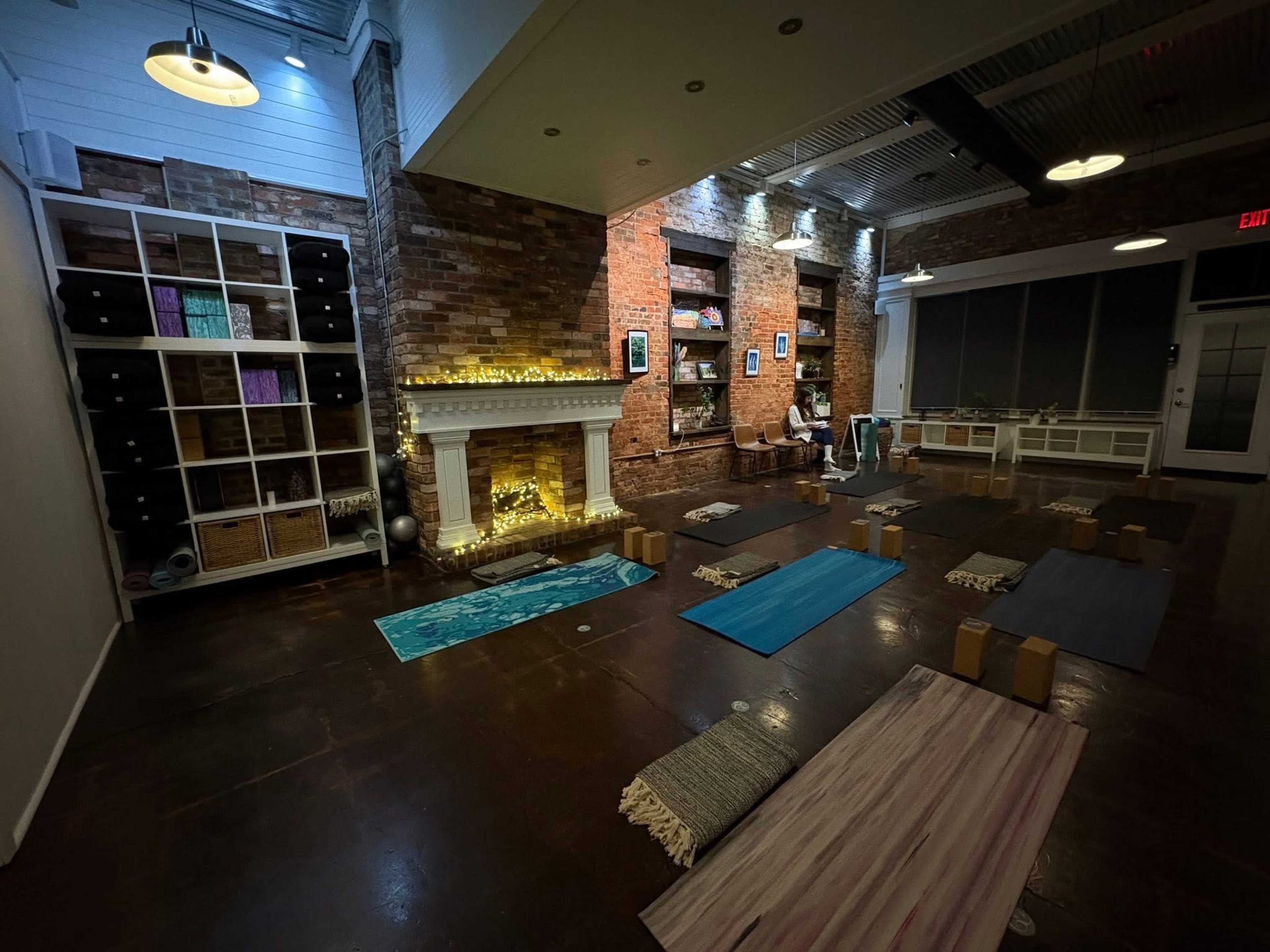 The image shows a dimly lit yoga studio with several yoga mats arranged on the floor, a decorative fireplace with soft lighting, and shelves filled with various items along the walls.