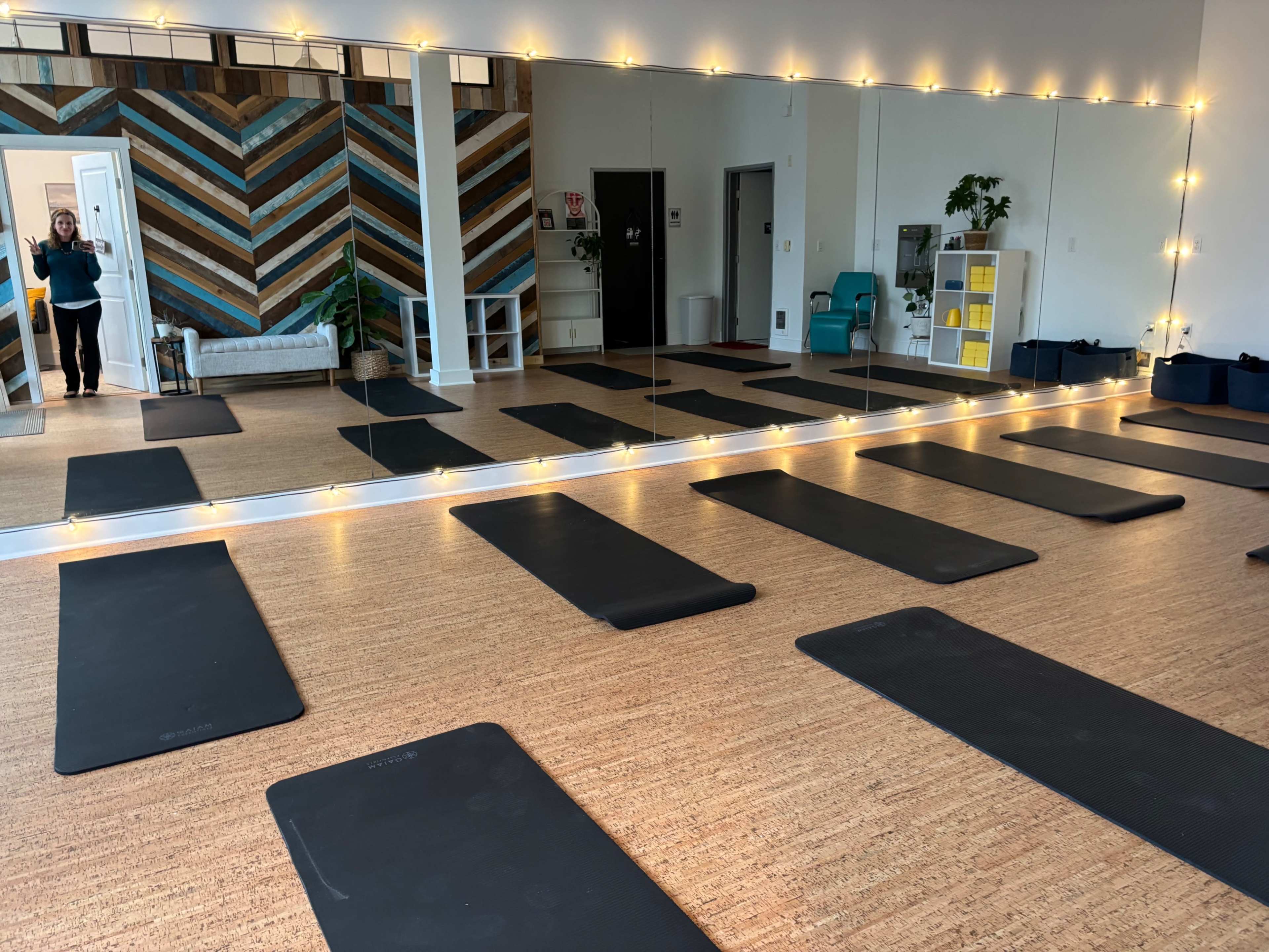 The image shows a yoga studio with multiple black mats arranged on a wooden floor, reflecting a colorful wooden accent wall and soft lighting.