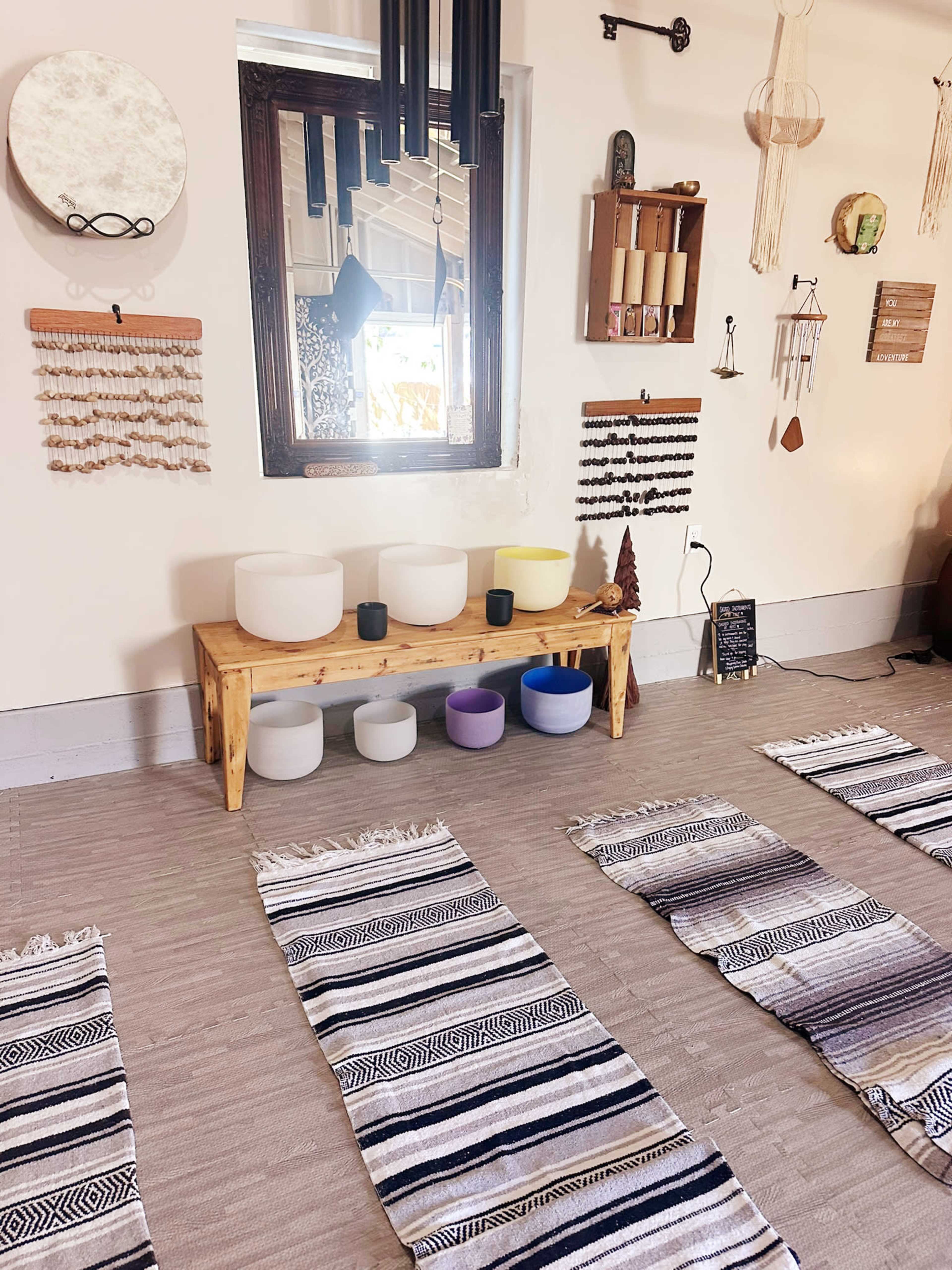 The image shows a serene room designed for meditation or relaxation, featuring a bench with various sound bowls, decorative wall hangings, and patterned mats arranged on the floor.