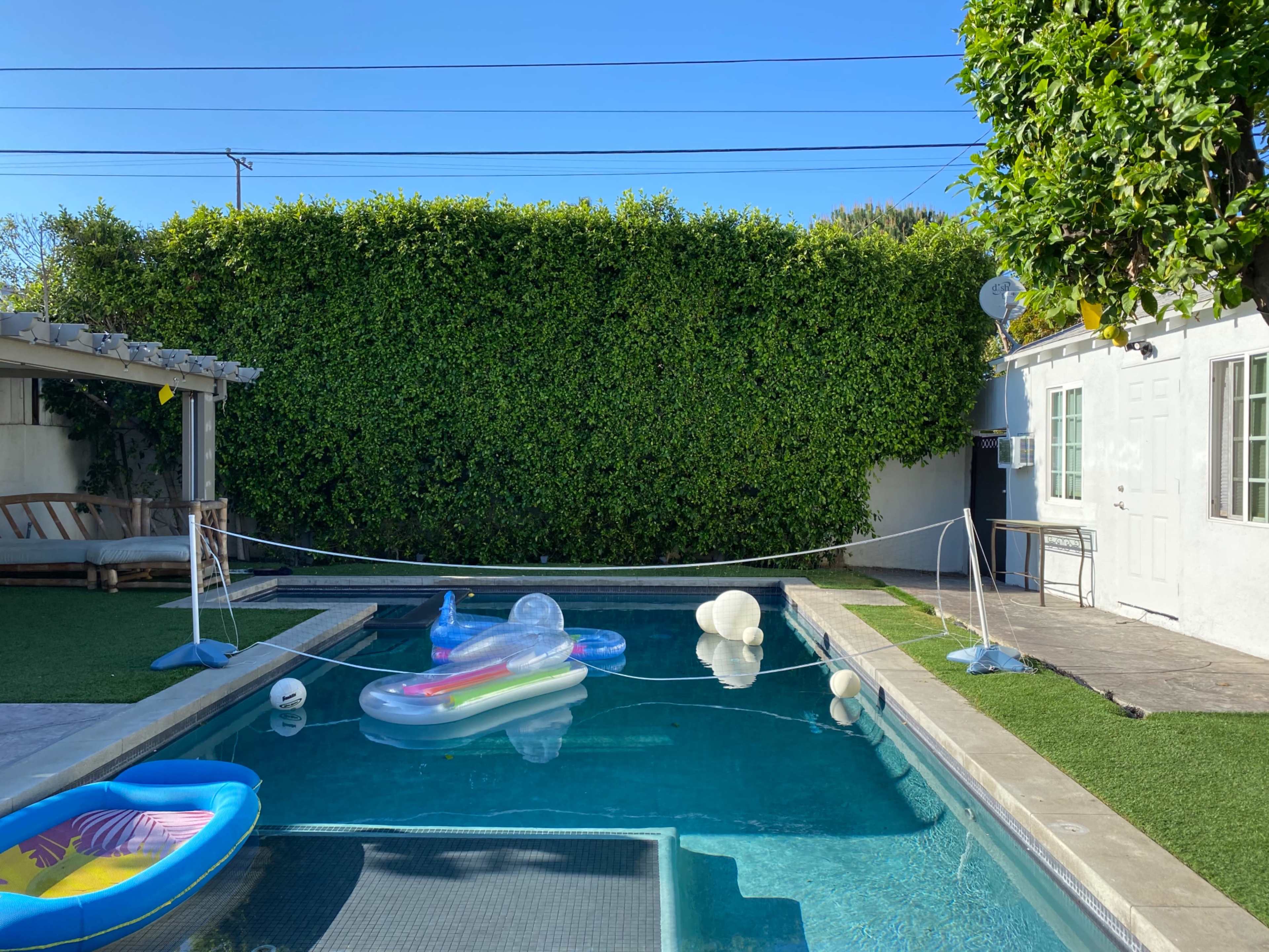 A clean swimming pool surrounded by greenery, with various inflatable floats and a white building to the right.