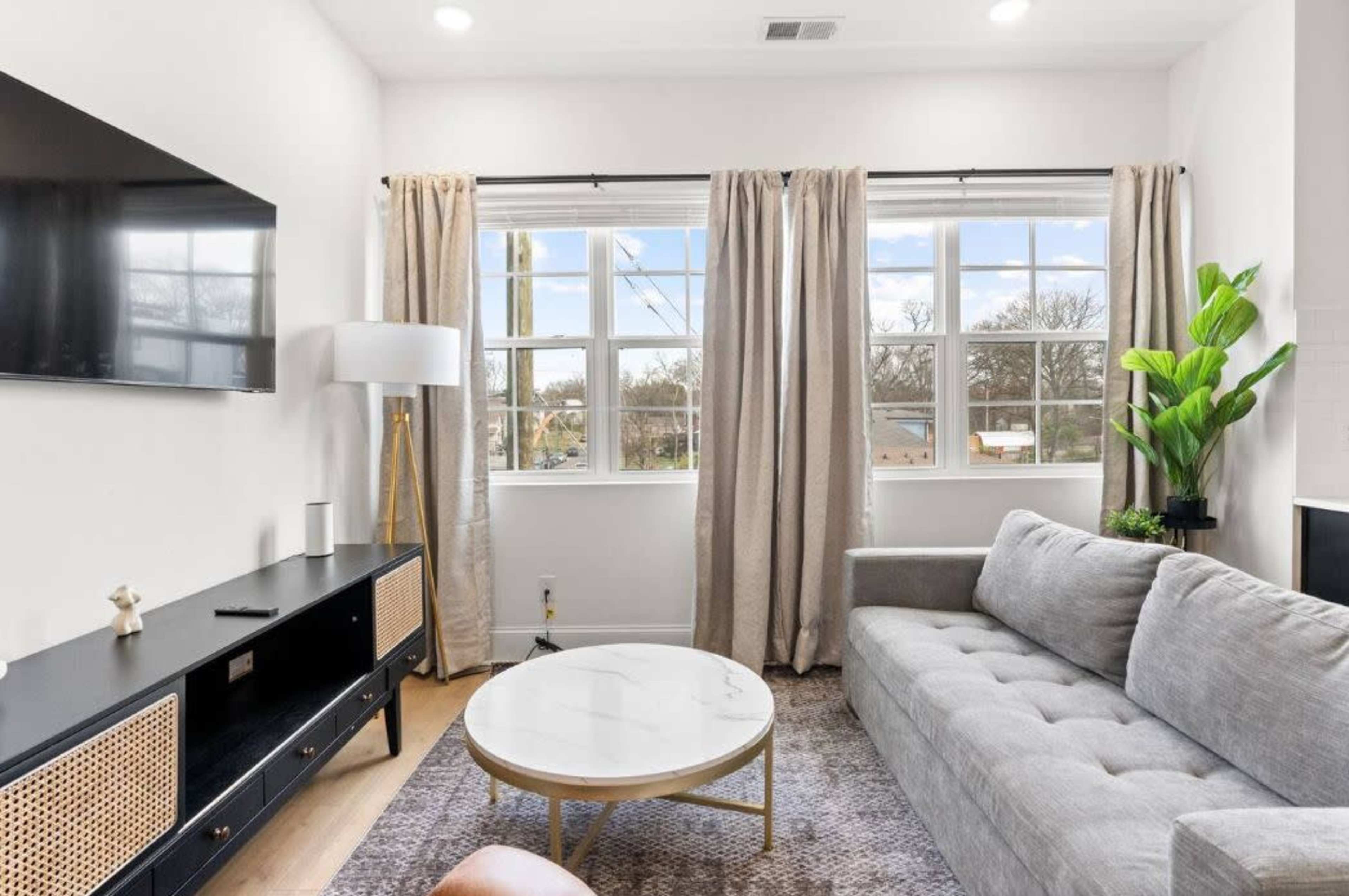 A modern living room features a gray sofa, a round coffee table, a television on the wall, and large windows with curtains letting in natural light.