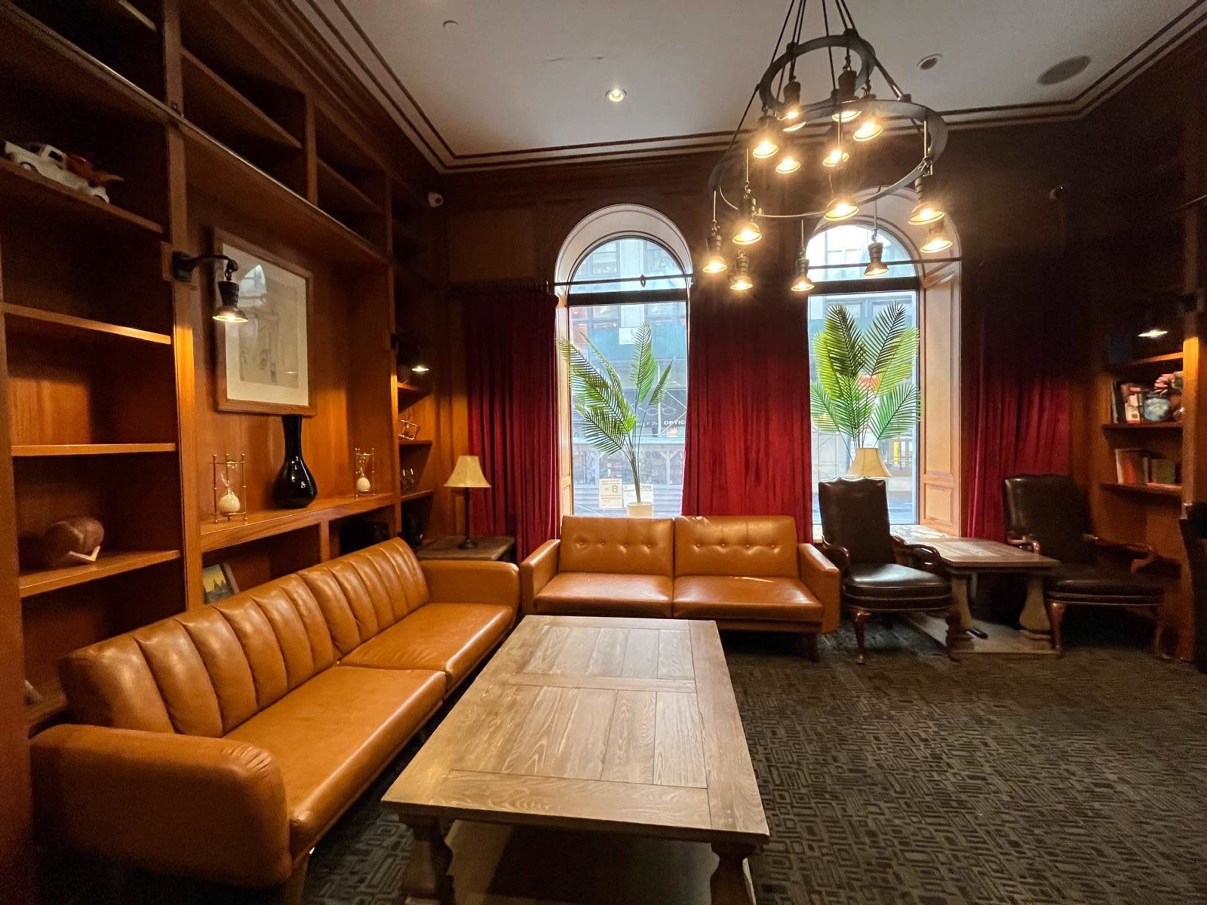 A well-furnished room featuring a brown leather sofa, a wood table, and large windows with red curtains and palm plants outside.