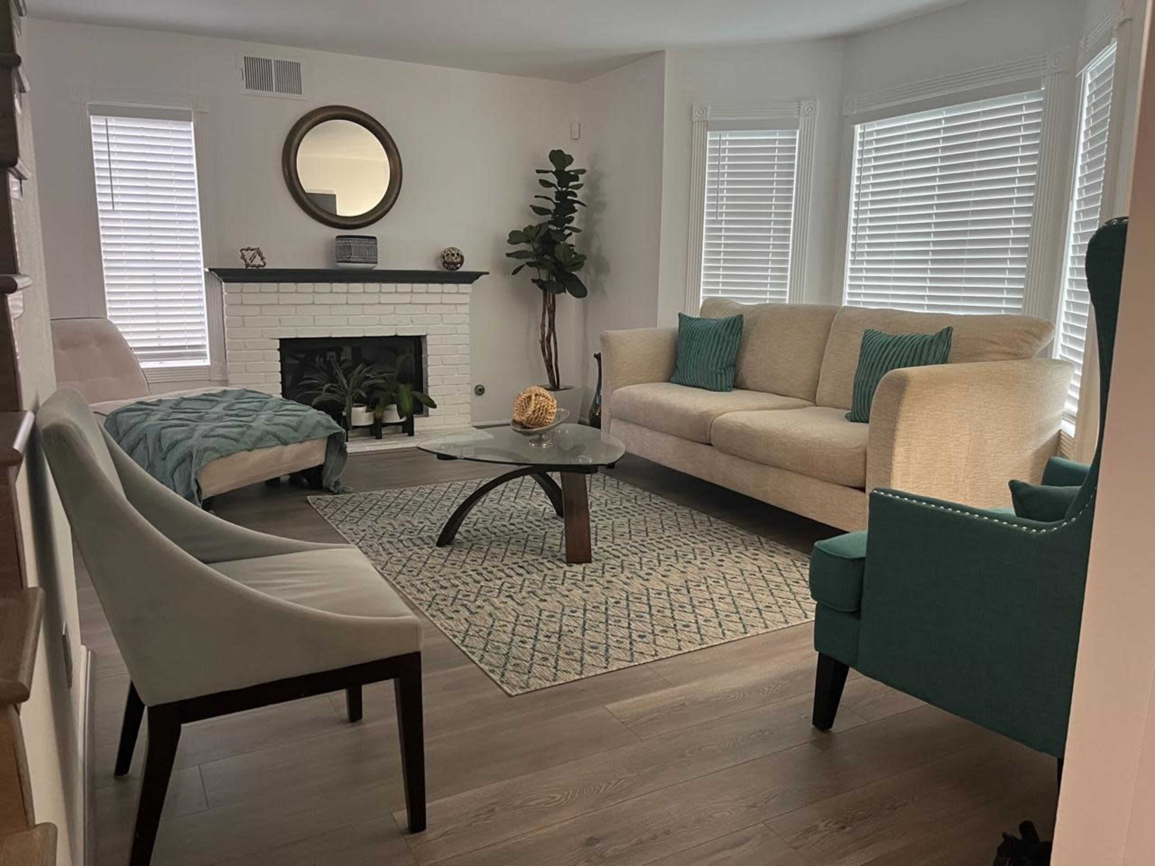 The living room features a light-colored sofa, a glass coffee table, a green armchair, and a decorative area rug, with large windows allowing natural light to fill the space.