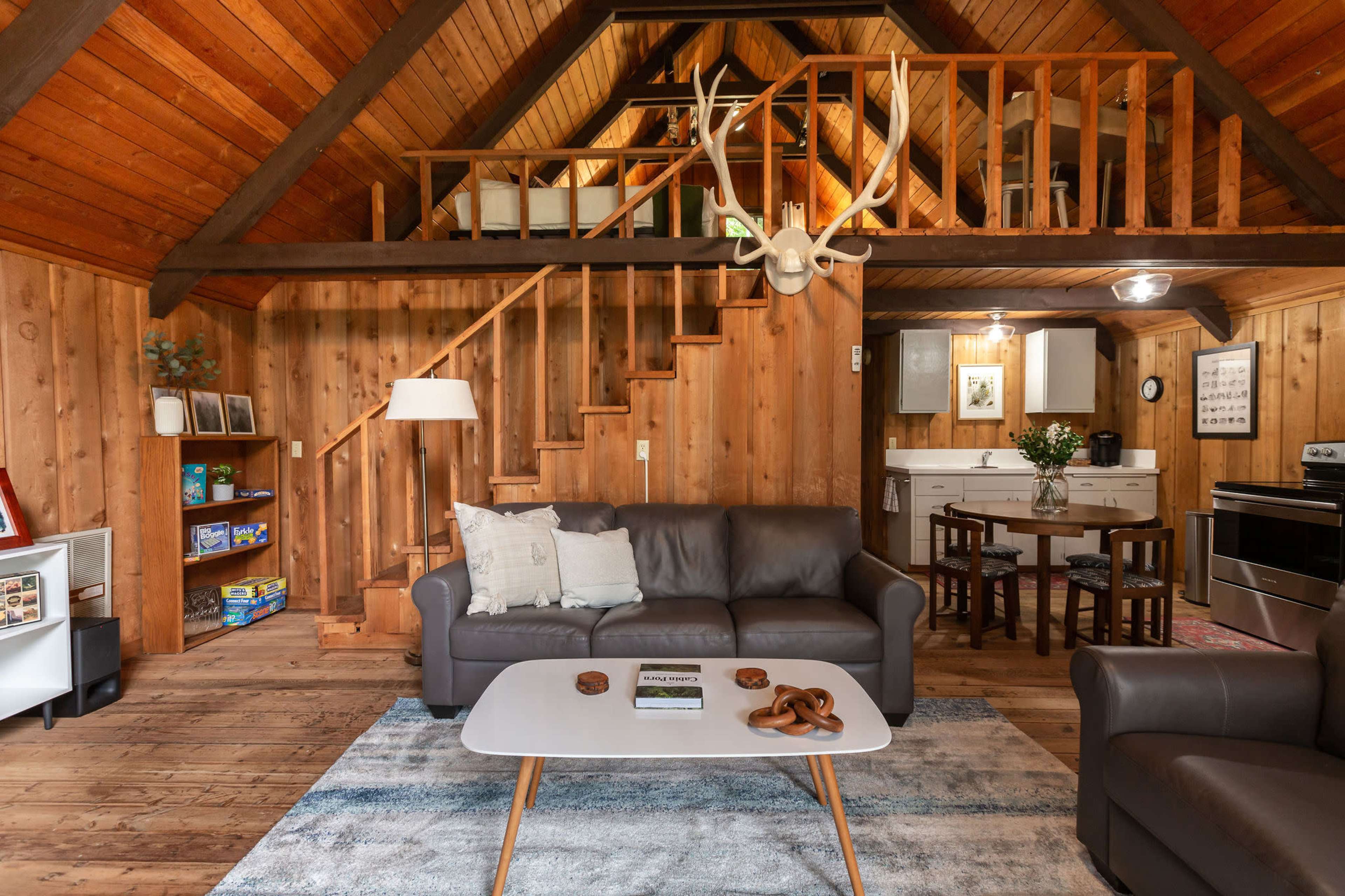 A cozy wooden interior features a living area with a sofa, coffee table, and a staircase leading to a loft, with a dining area and kitchen visible in the background.
