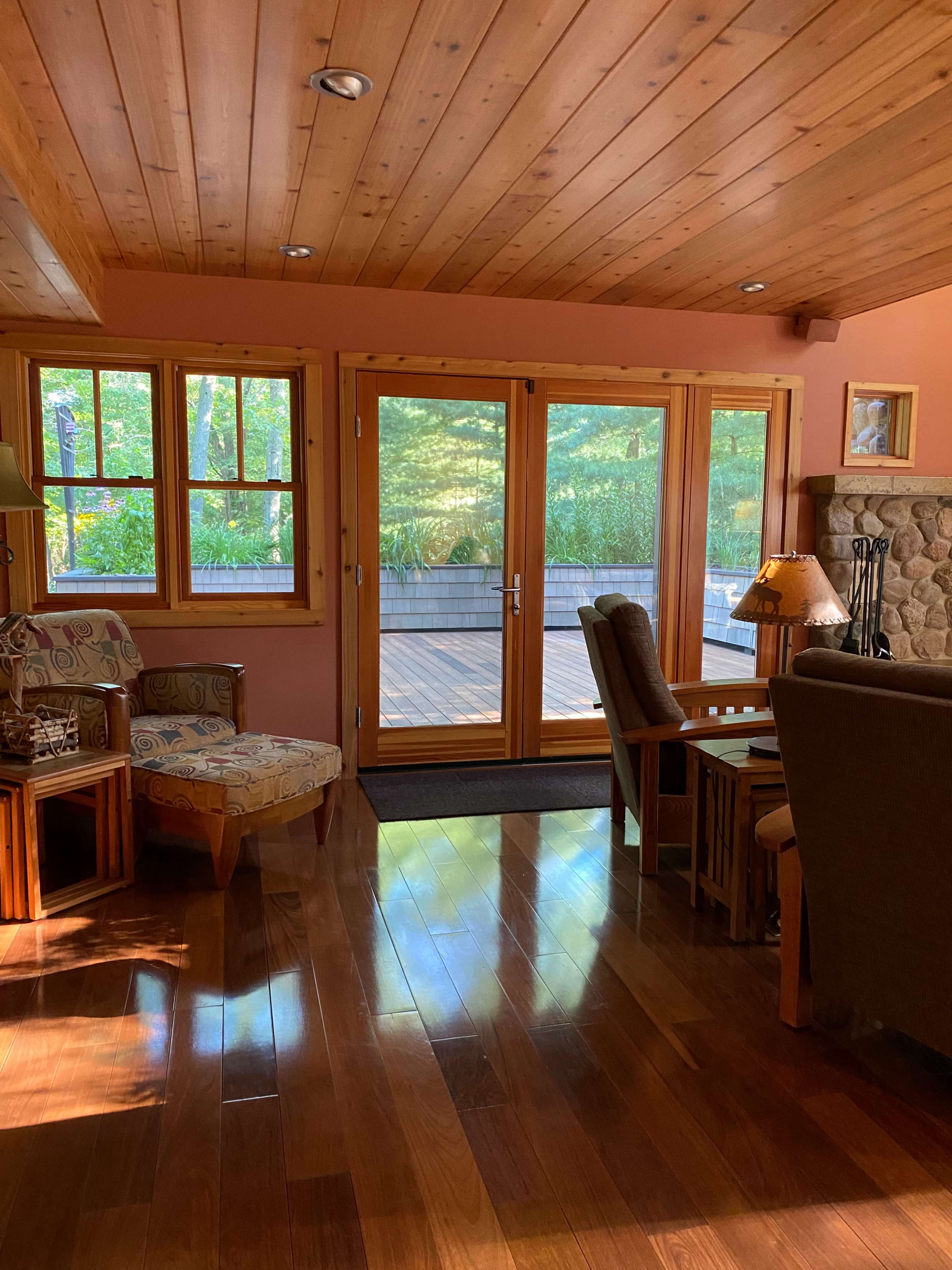 The image shows a wooden-floored living room with large glass doors leading to an outdoor deck, surrounded by trees.