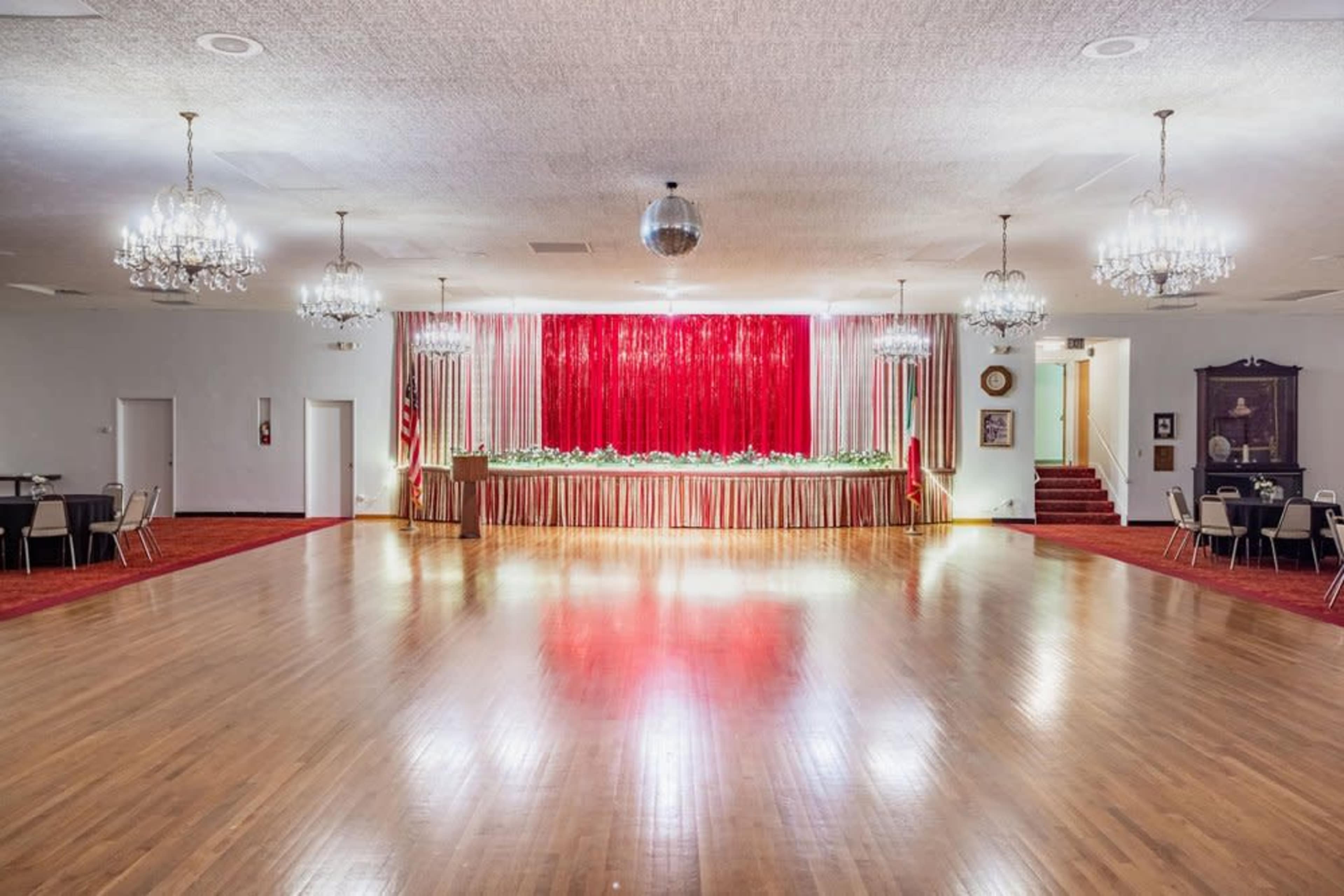 The image shows a spacious event hall with a polished wooden floor, adorned with a stage at the front featuring red curtains and decorative greenery.
