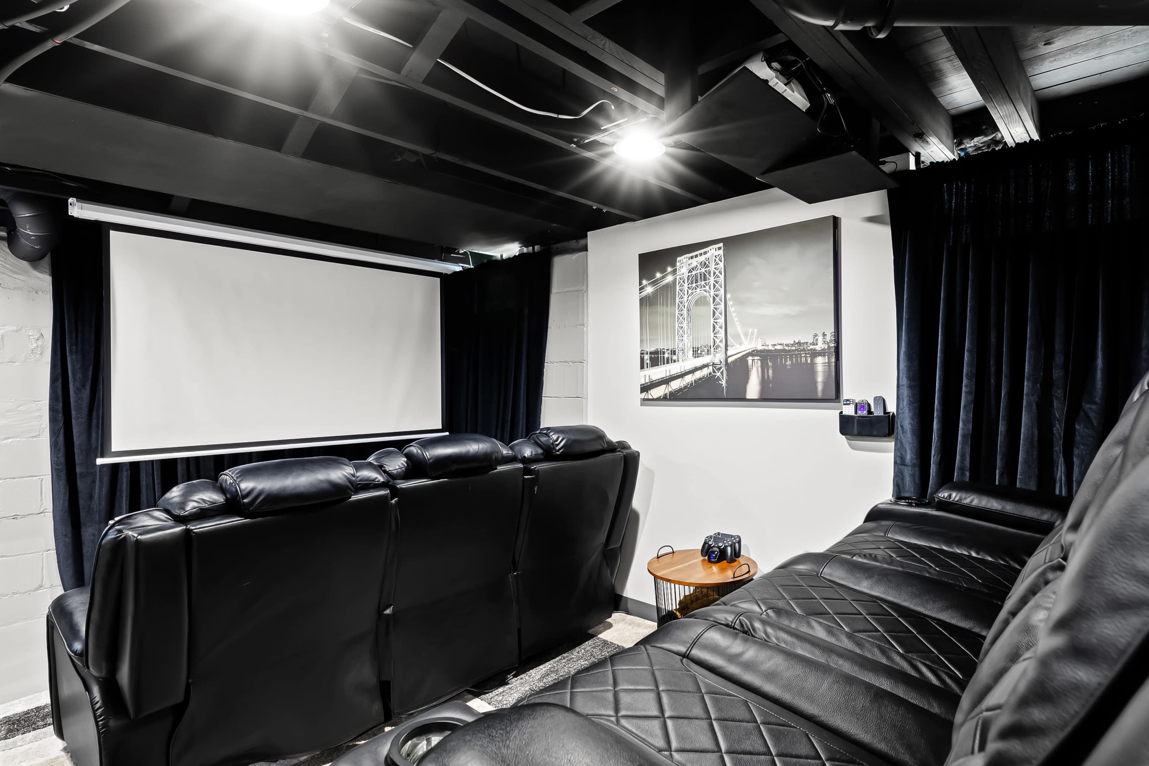 A darkened home theater room with black leather seating, a projector screen, and a wall-mounted photograph of a bridge.