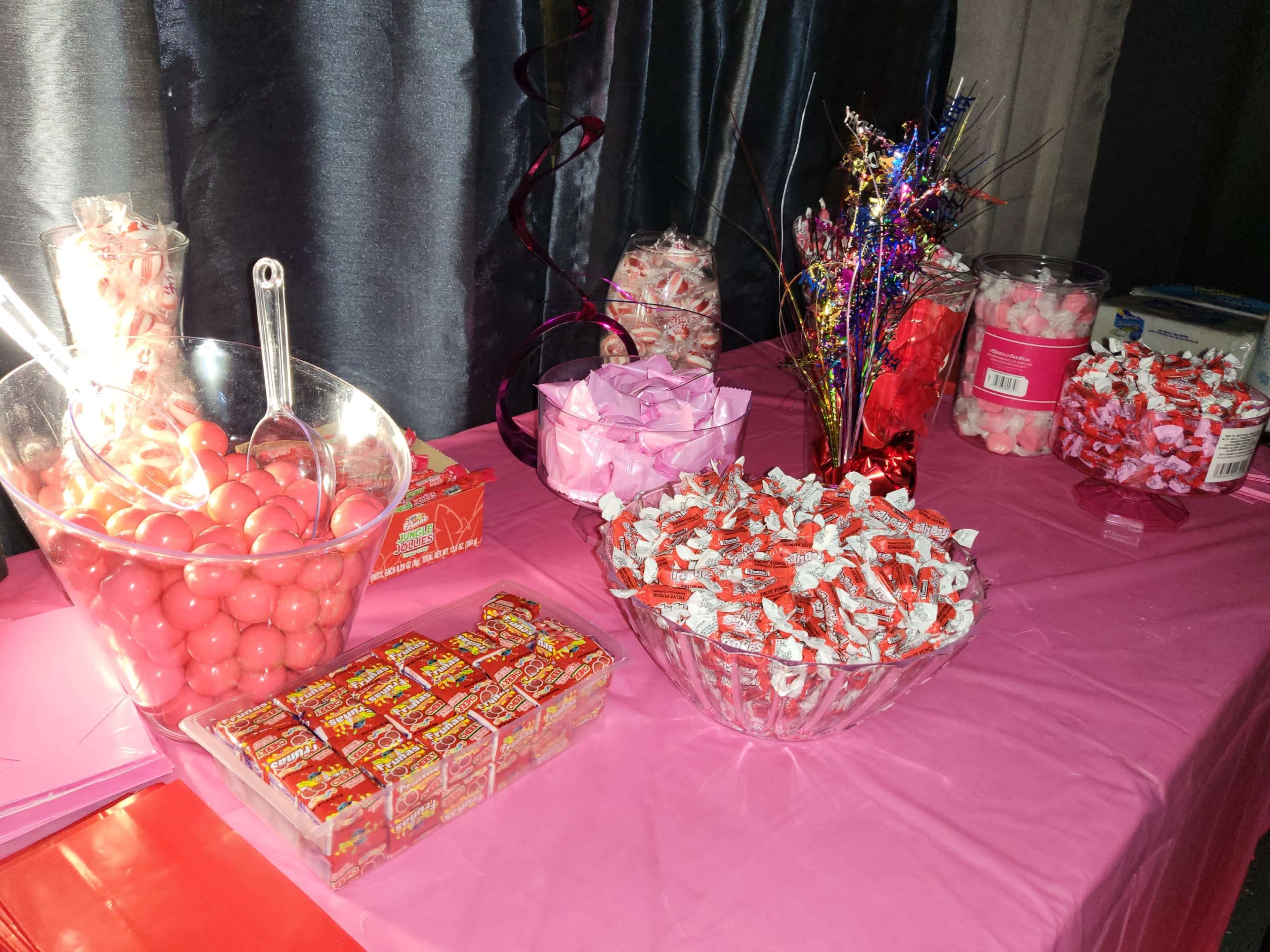 A table covered in pink cloth displays various containers filled with colorful candies, including pink gumballs, festive decorations, and assorted wrapped treats.