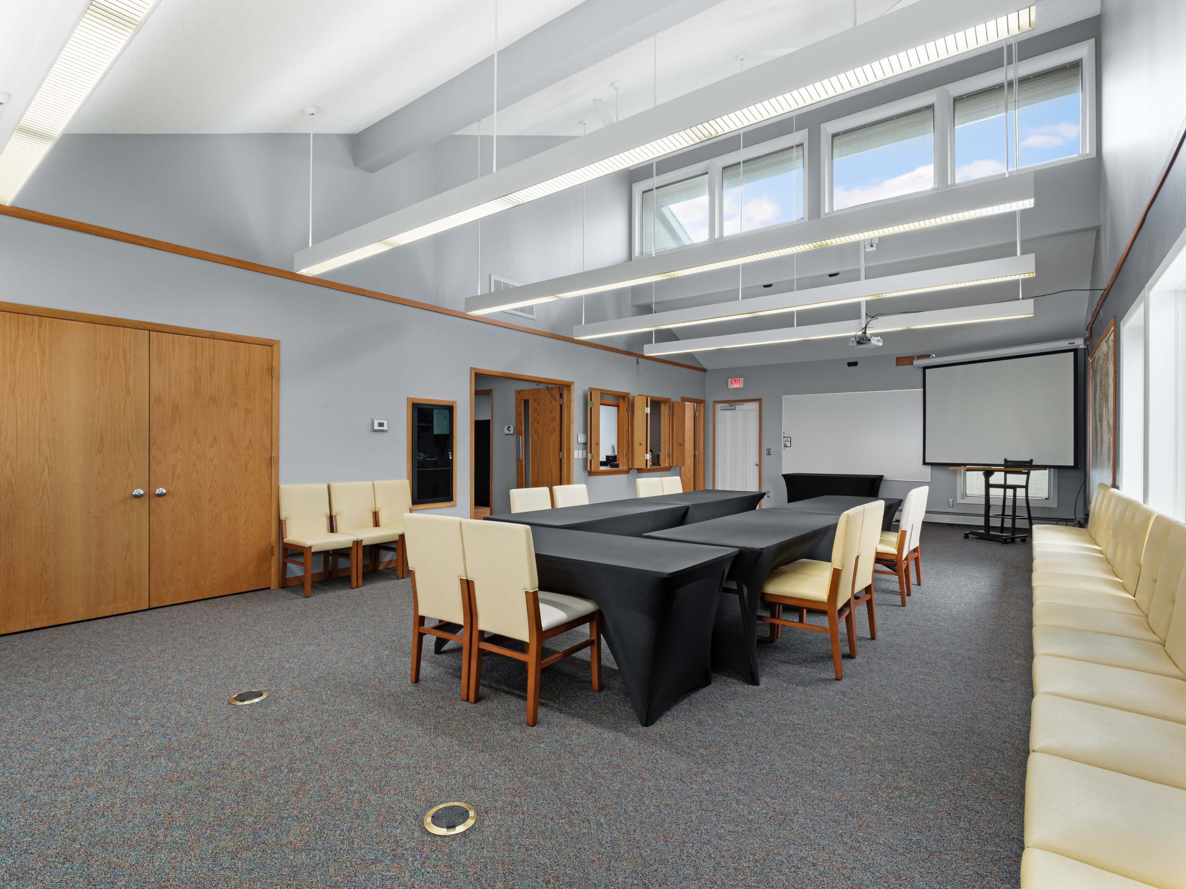 A conference room features a large black table surrounded by white chairs, with additional seating along the walls and large windows allowing natural light.