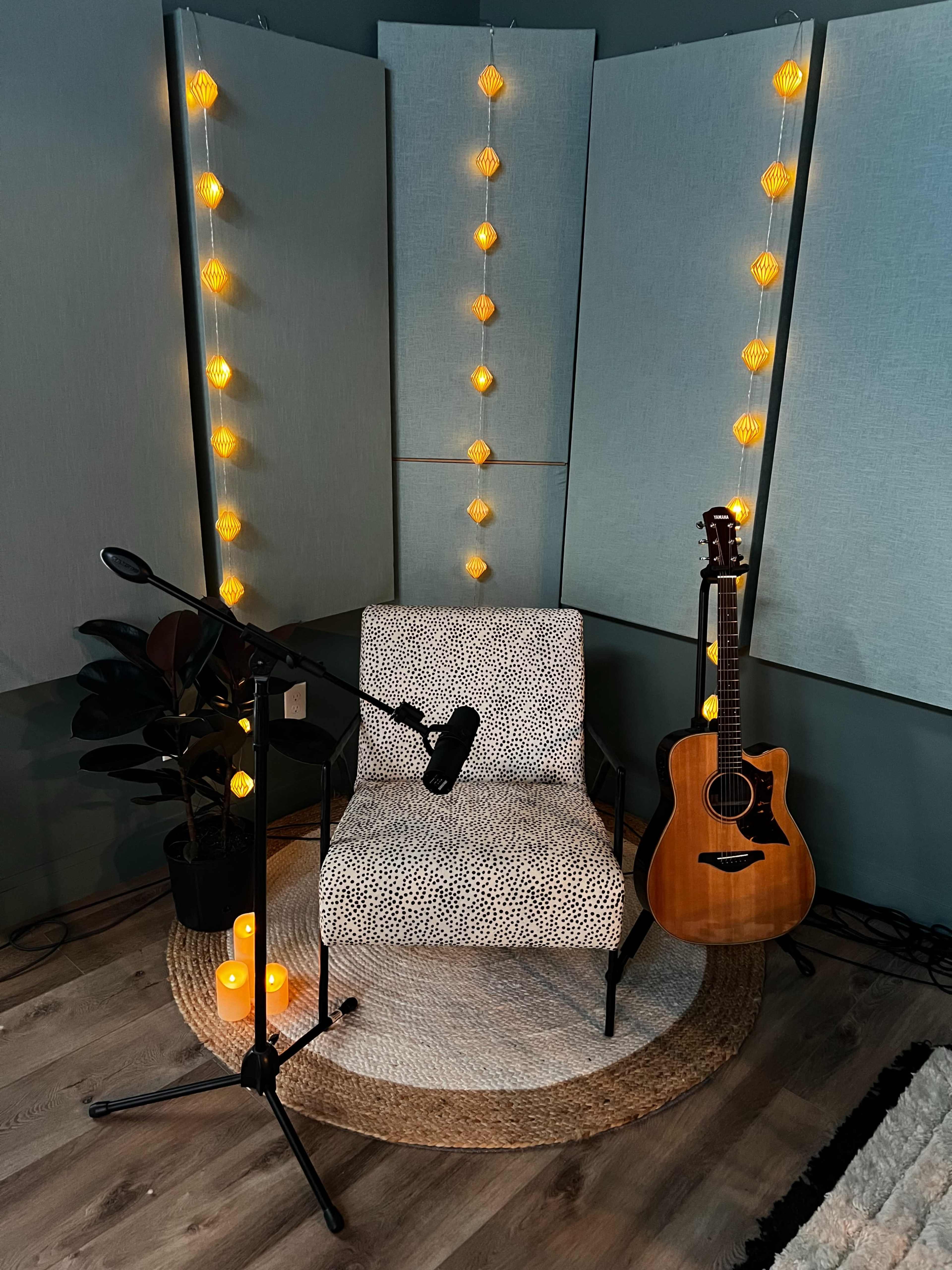 The image shows a cozy music setup featuring a patterned chair, a microphone on a stand, an acoustic guitar, warm string lights, and decorative plants in a softly lit room.