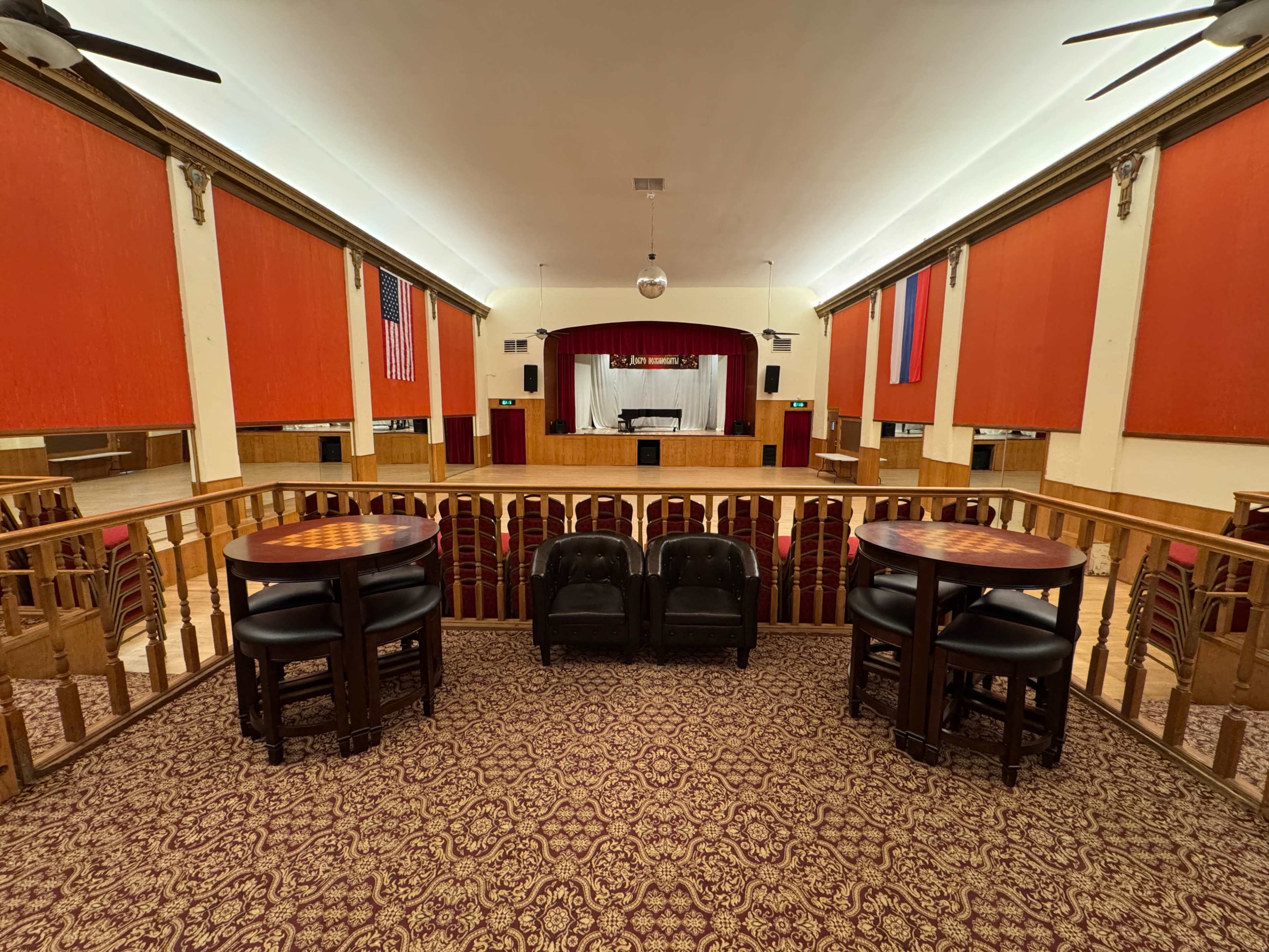 The image shows a spacious theater interior with a stage at the front, flanked by red curtains and two American flags, and features seating arrangements and a balcony overlooking empty chairs below.