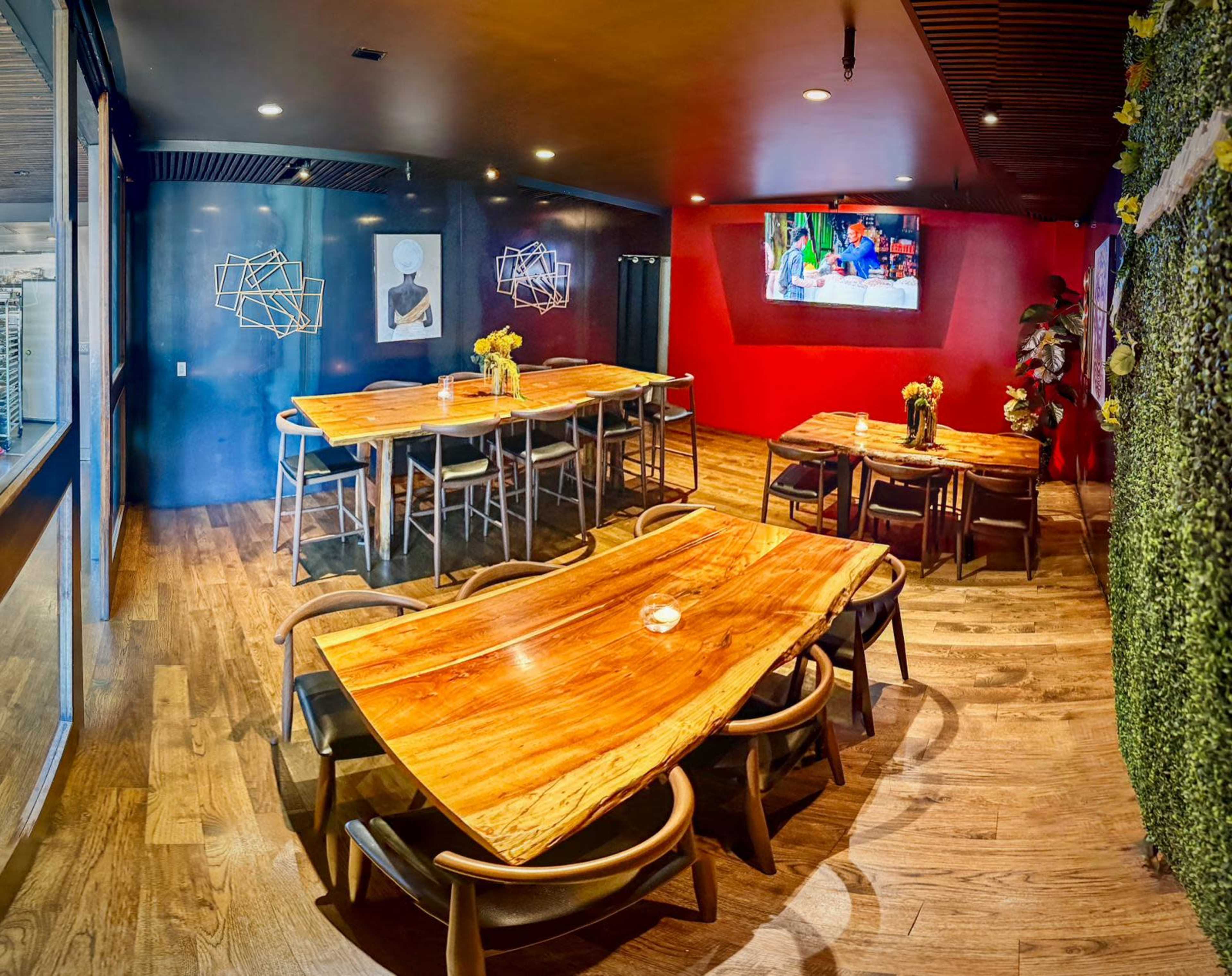 The image shows a modern dining area with several wooden tables and chairs, featuring textured walls in red and black, and decorative plants.