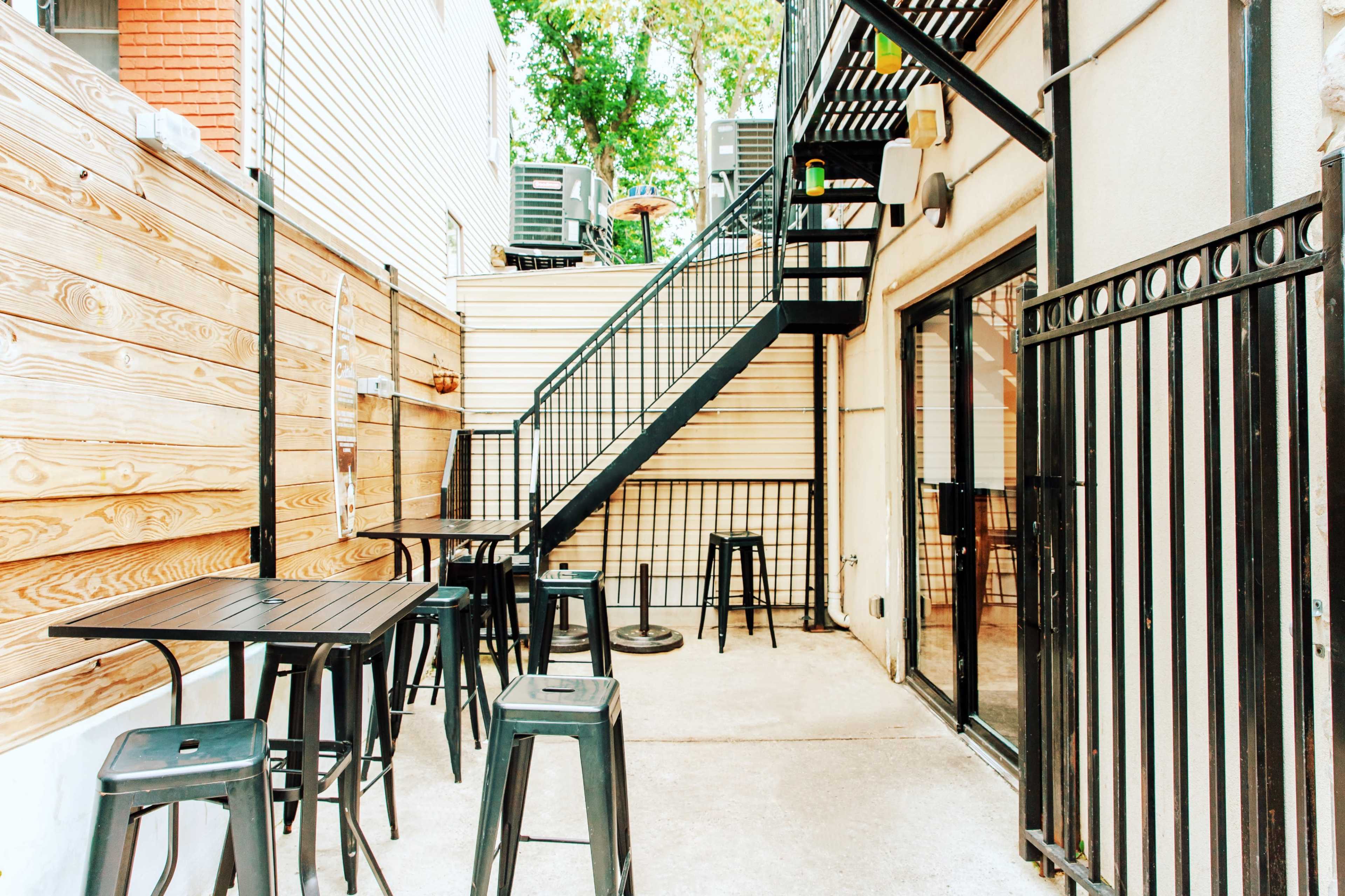 The image shows a small outdoor seating area with black metal tables and stools next to a staircase and wooden fences.