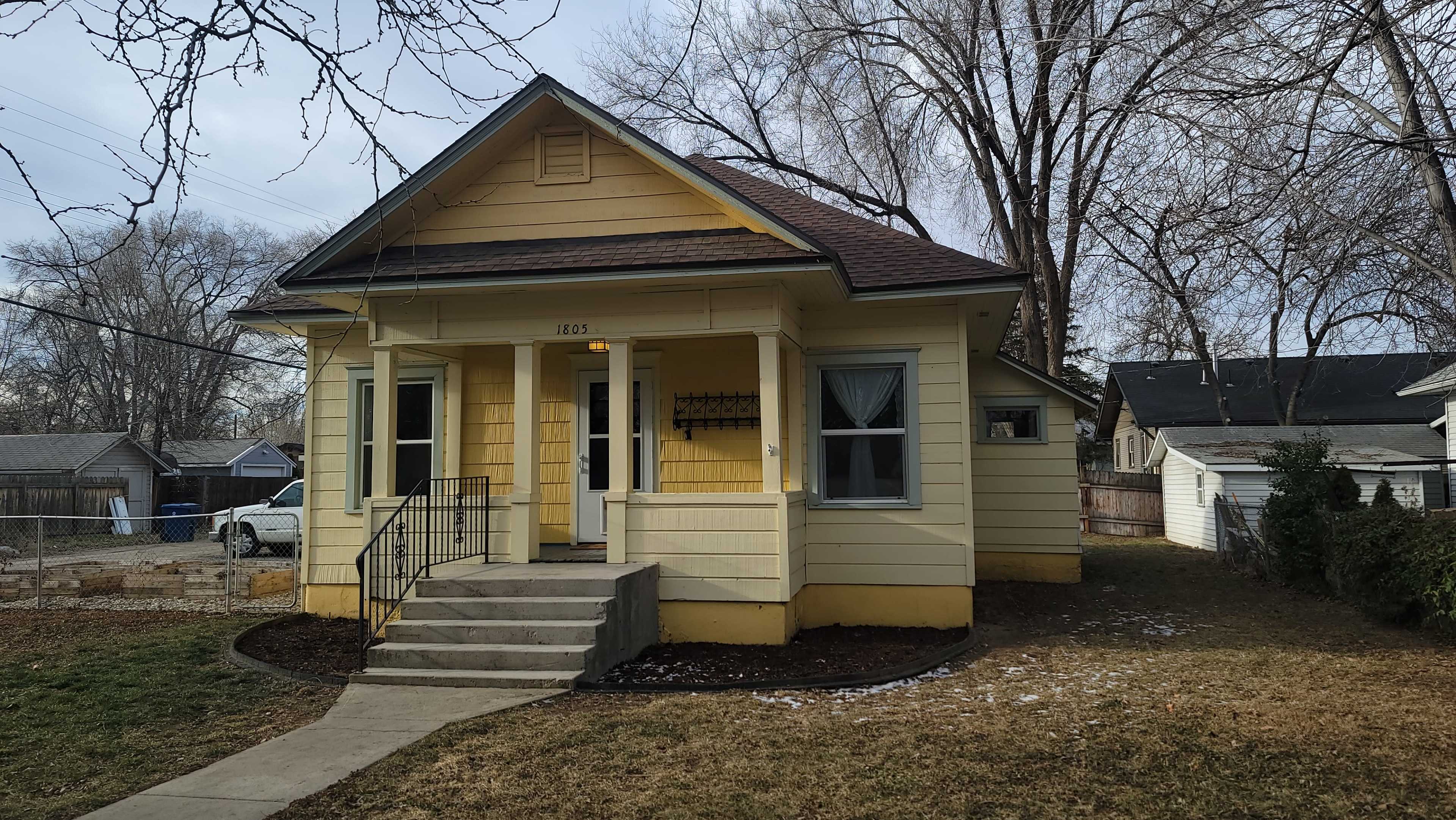 A yellow, single-story house with a front porch, stairs leading up to the entrance, and a yard featuring sparse grass and trees in the background.