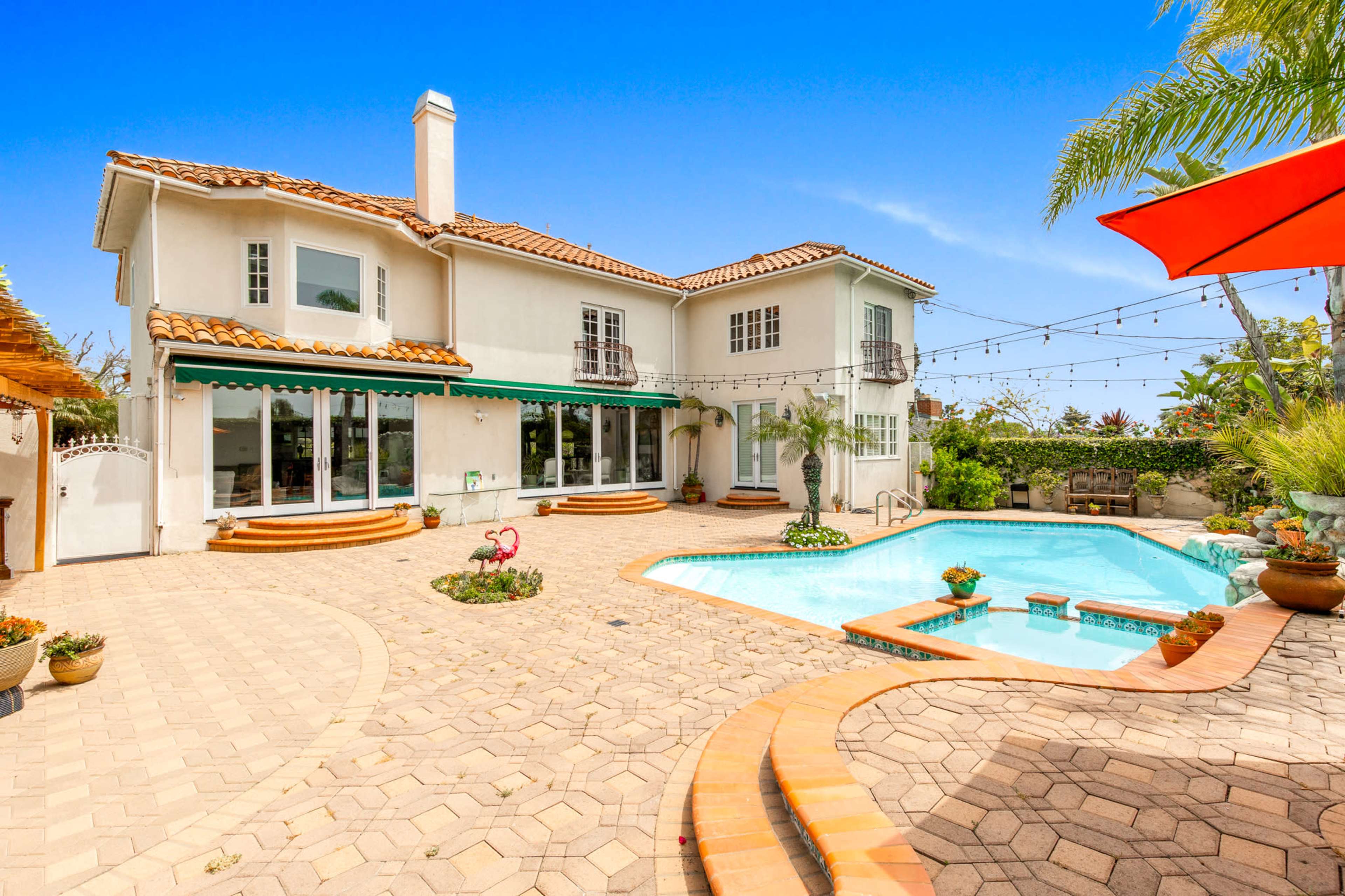 A two-story house with a red tile roof and a patio surrounds a swimming pool, flanked by potted plants and decorative features.