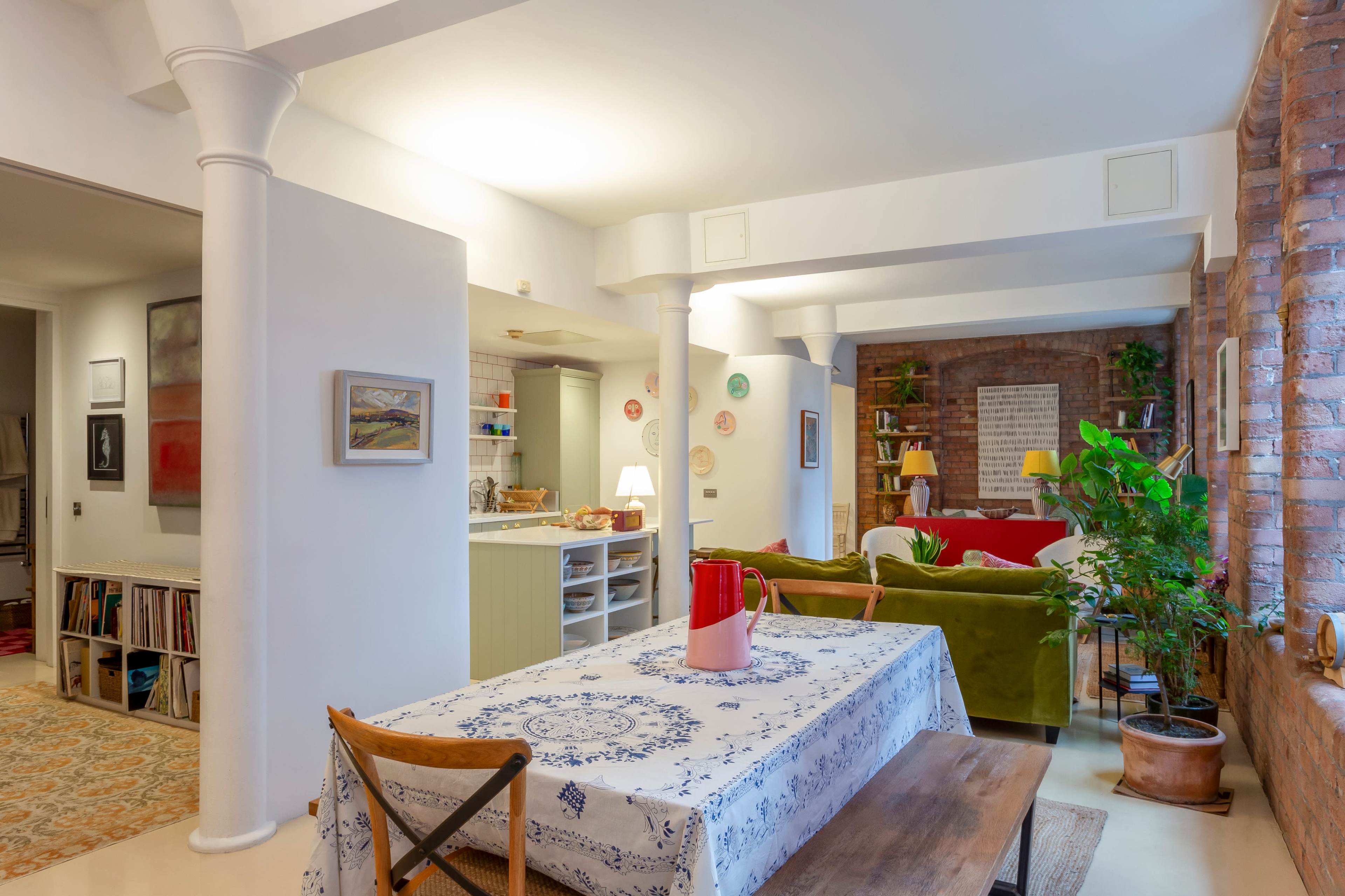 An open living space featuring a dining table with a patterned tablecloth, a green sofa, and a kitchen area in the background.