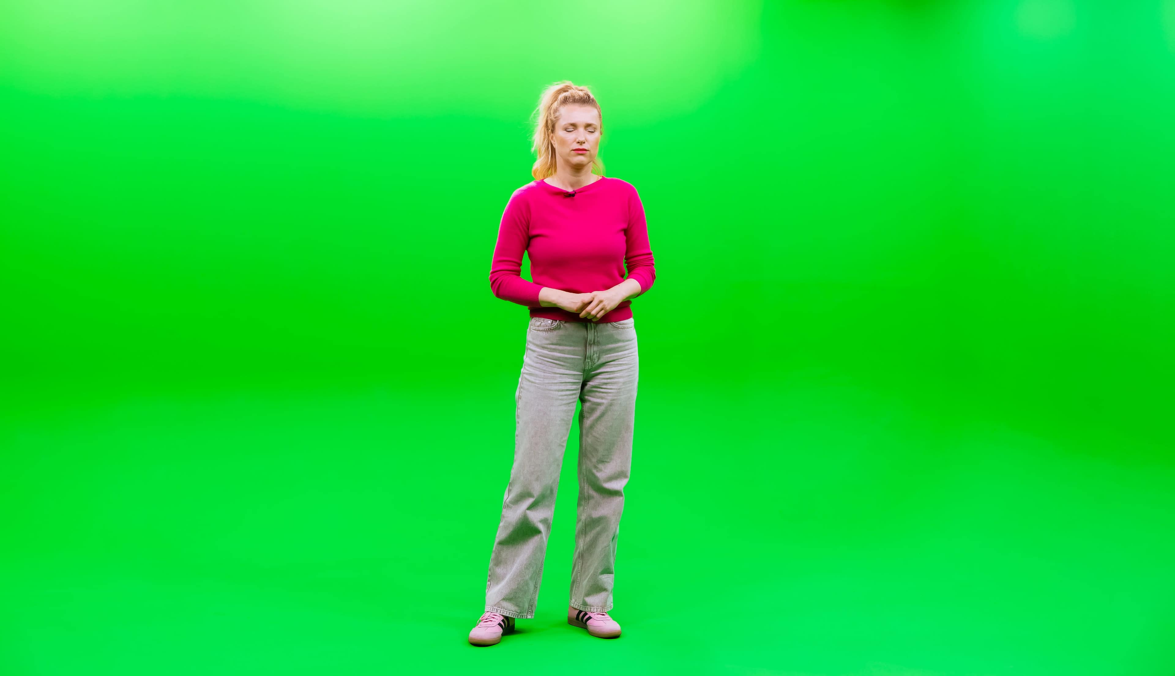 A person stands in front of a bright green background, wearing a pink long-sleeve shirt and gray pants.