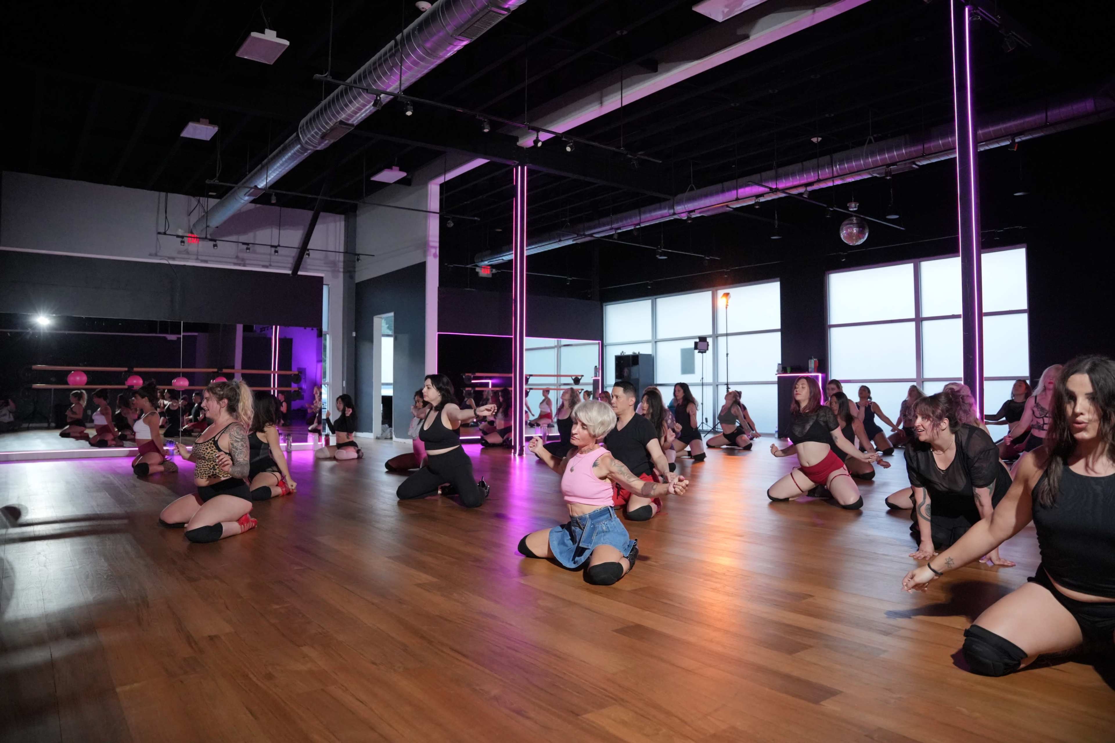A dance studio filled with performers practicing choreography on a polished wooden floor, with purple lighting illuminating the space.