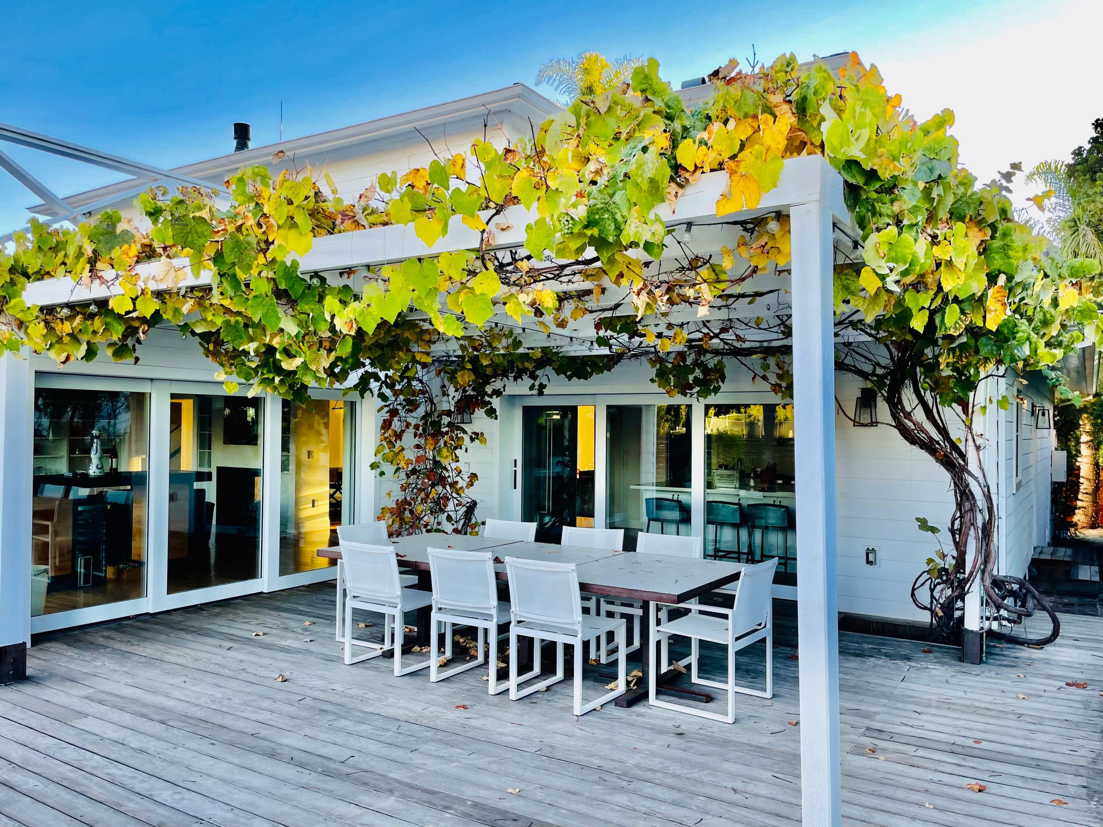A wooden deck with a dining table and chairs under a pergola covered with green and yellow foliage, adjacent to a house with large glass doors.