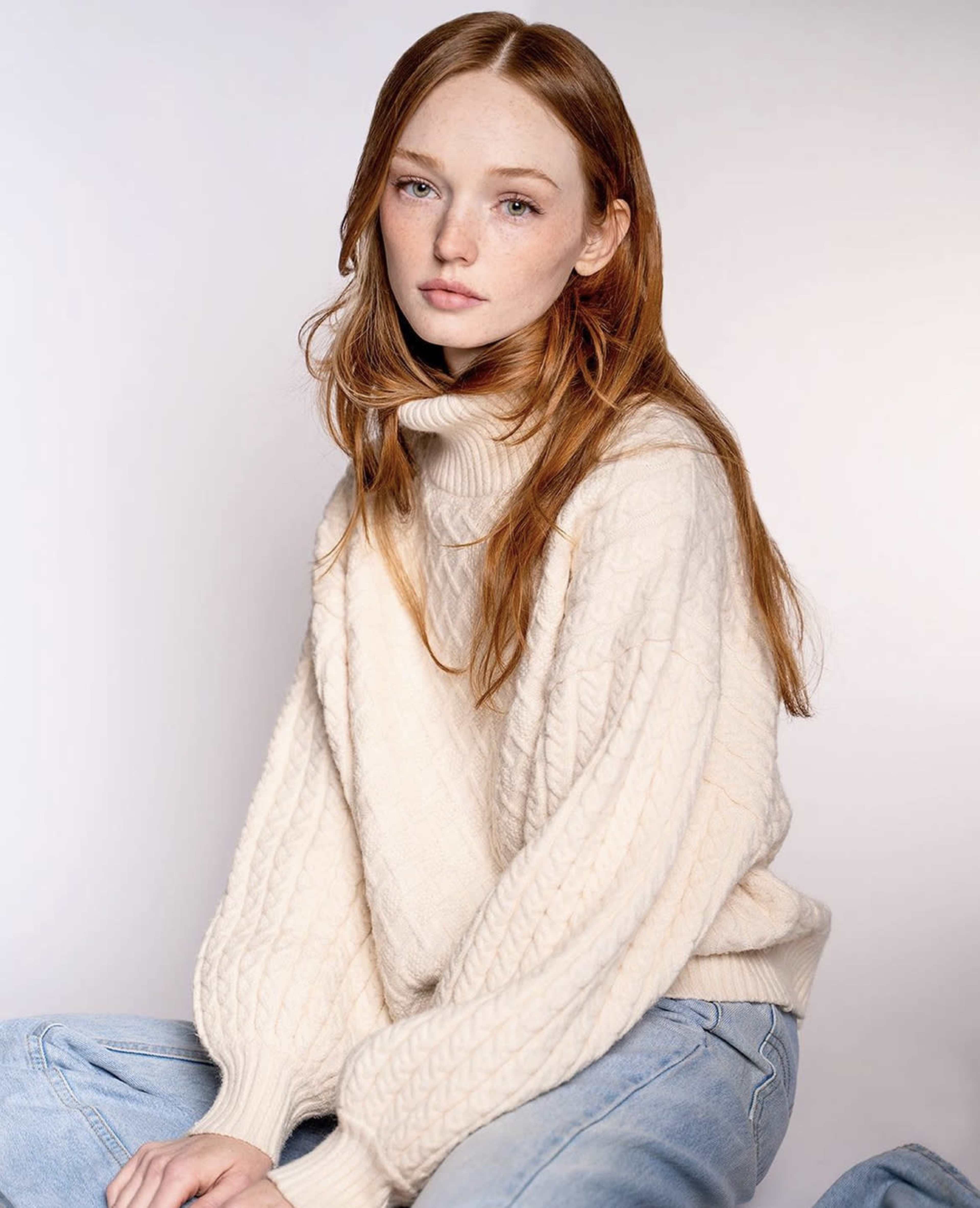 A young woman with long red hair sits against a neutral background, wearing a cream-colored turtleneck sweater and light blue jeans.
