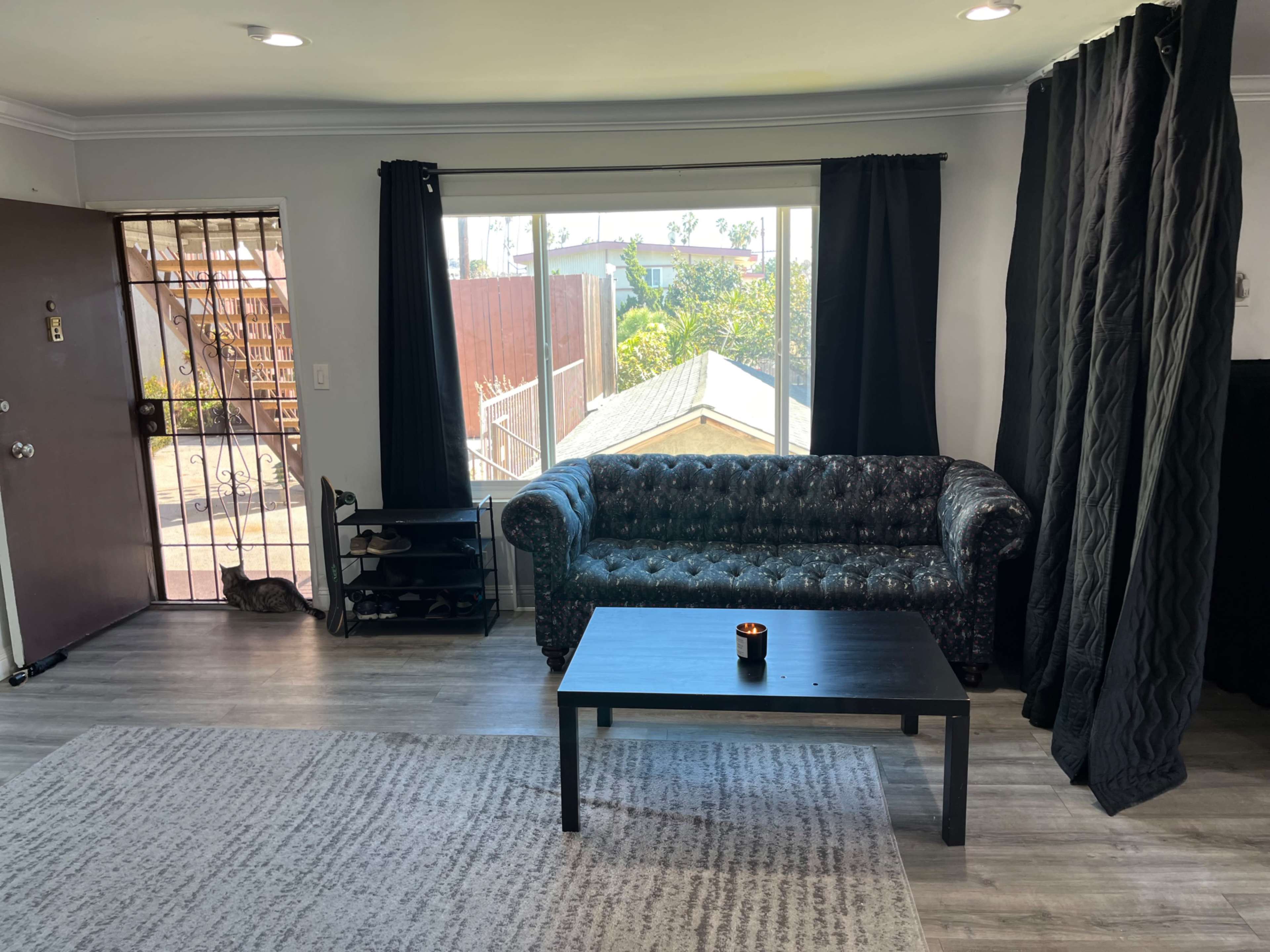 The living room features a black couch and a coffee table, with large windows providing natural light and a view of the outdoors.