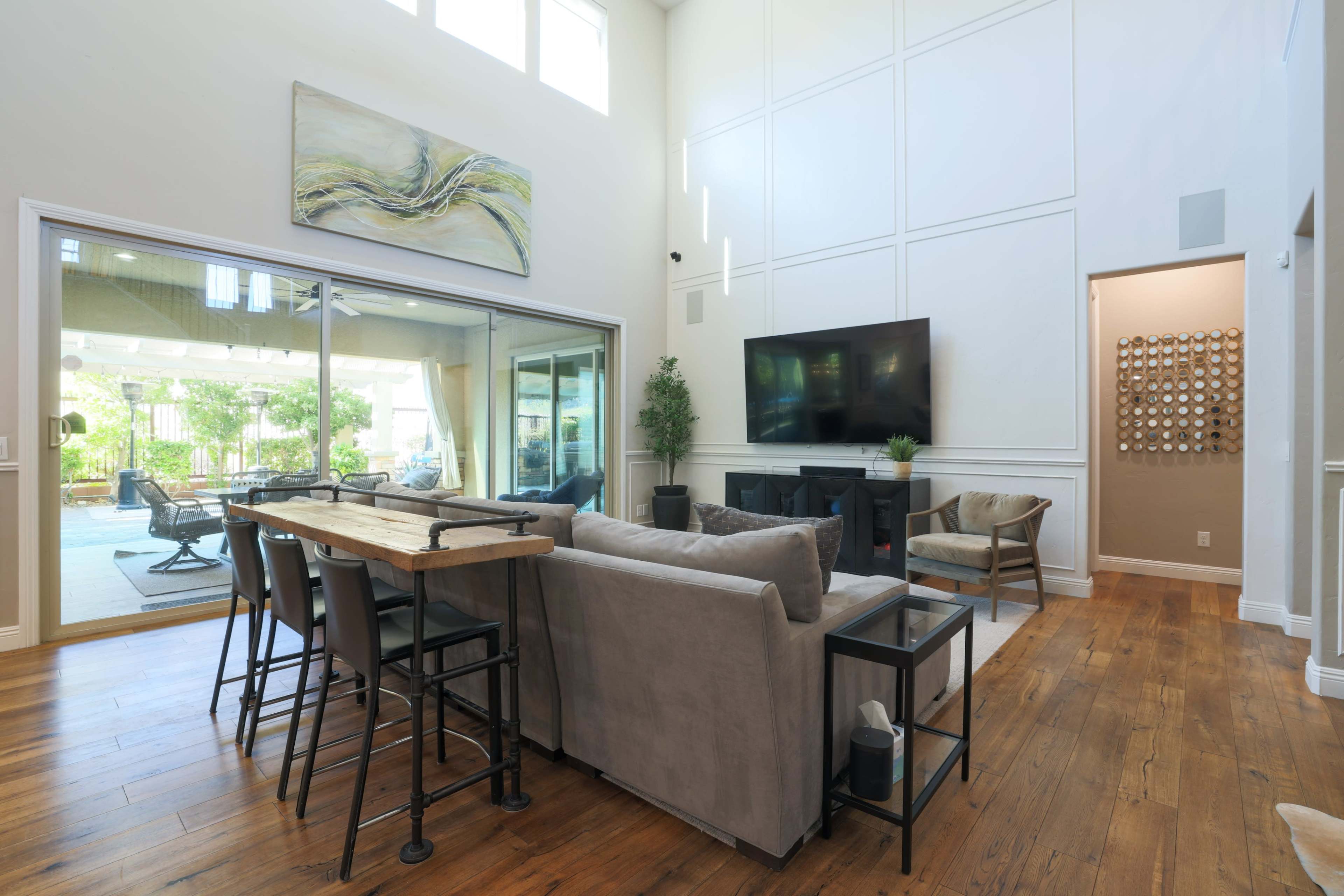 The image shows a spacious living room with large windows, a wooden table with bar stools, a flat-screen TV on the wall, and a view of an outdoor patio through sliding glass doors.