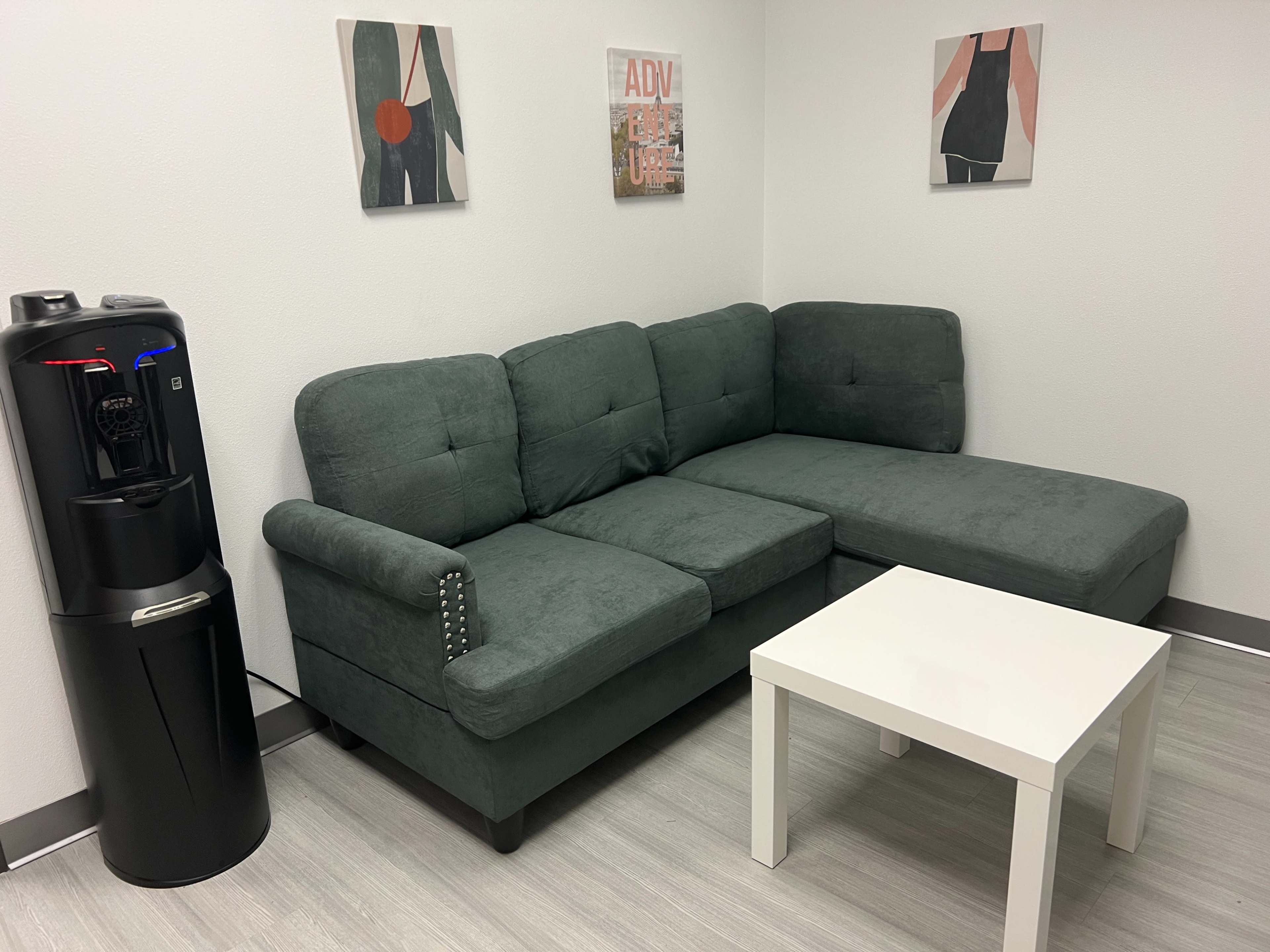 A small lounge area with a grey sectional sofa, a white coffee table, and a water cooler against a plain wall with two framed artworks.