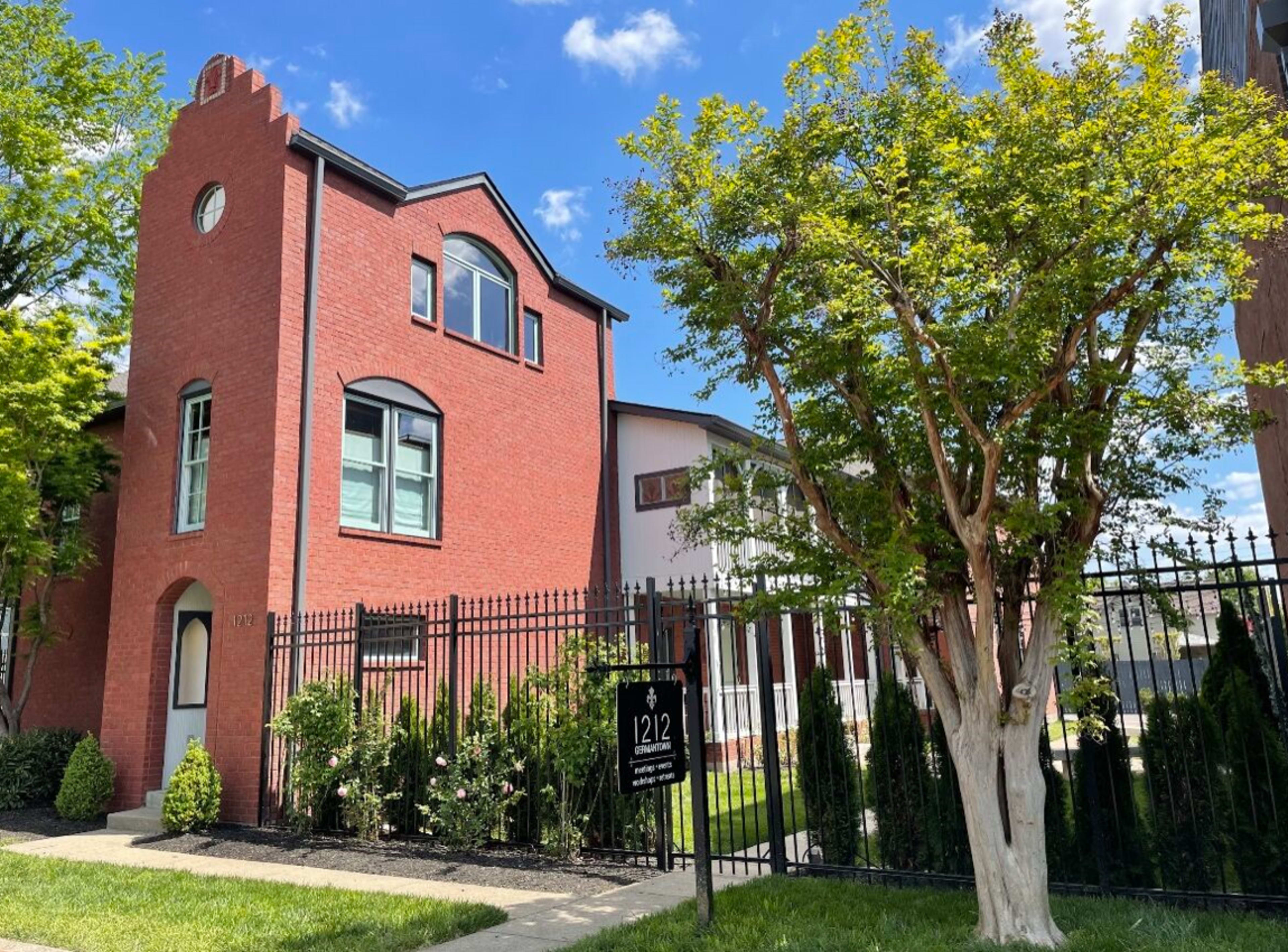 A red brick house with a modern architectural style is flanked by a large tree and a black iron fence.
