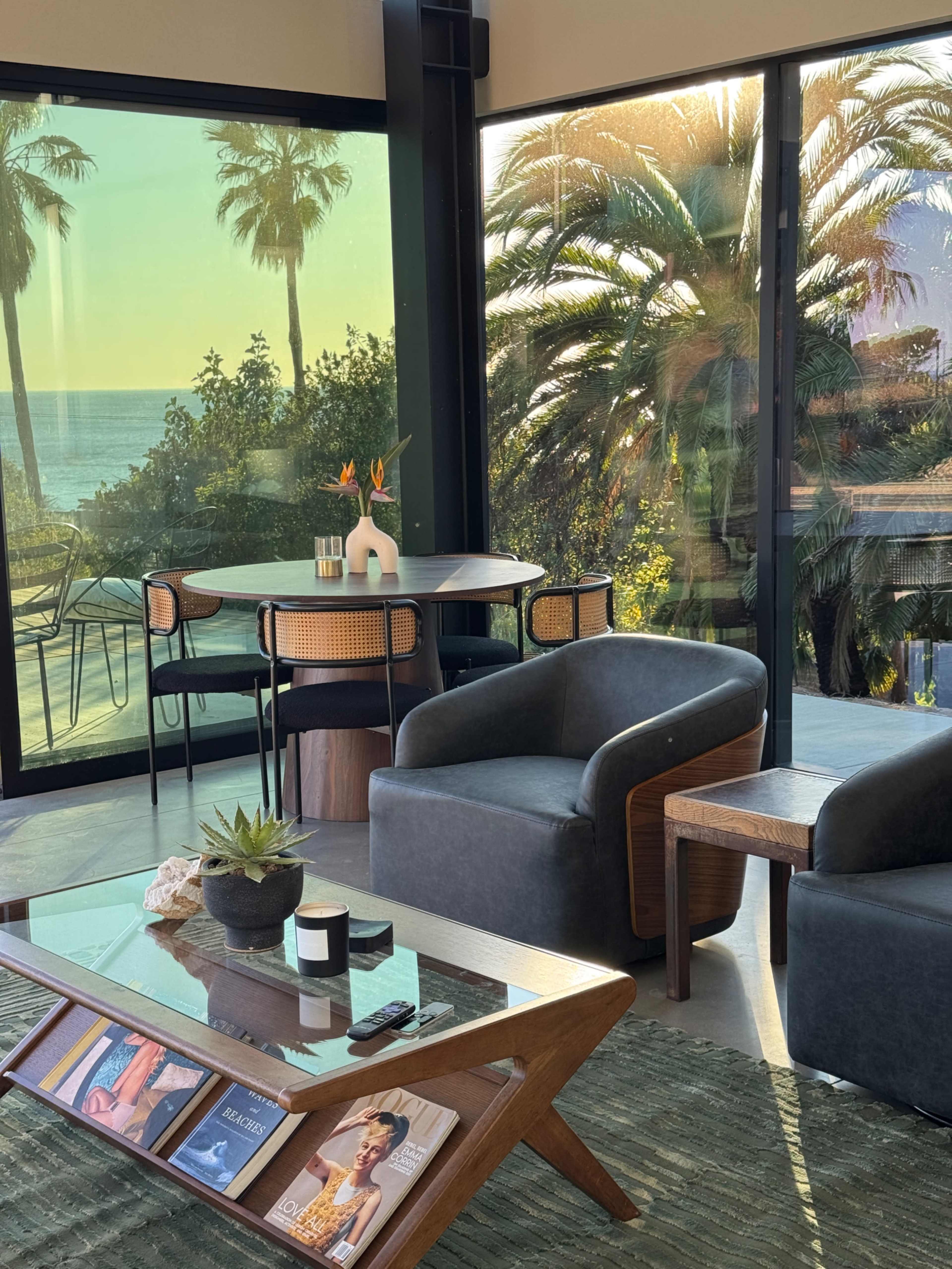 A modern living room features a glass wall overlooking palm trees and the ocean, with a dining table set for four and stylish seating.