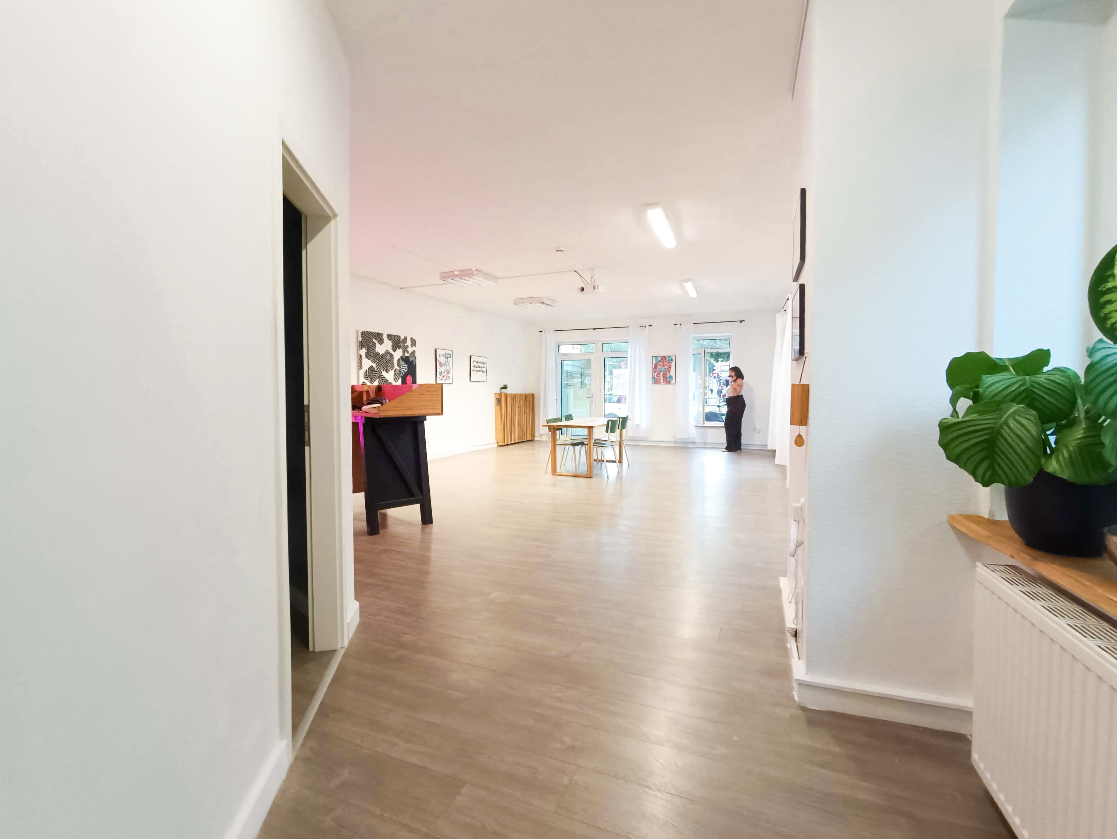 The image shows a bright, open hallway leading into a spacious room with wooden flooring and a few pieces of furniture, including a table and chairs.