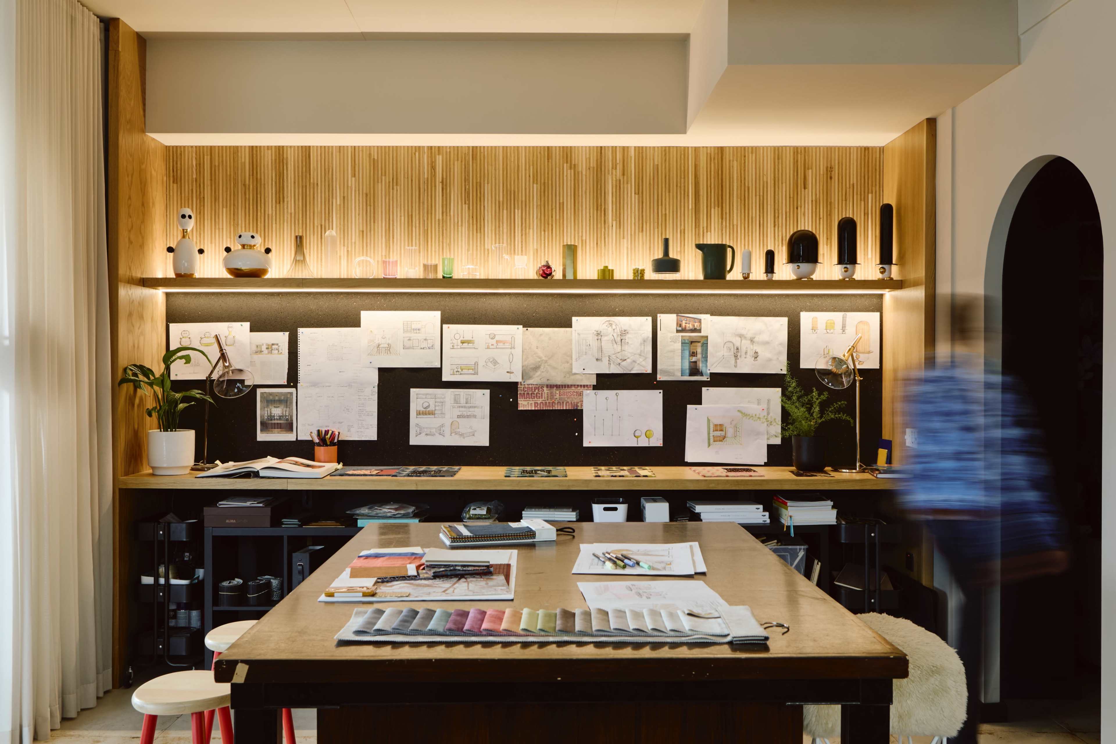 A creative workspace features a wooden table surrounded by sketches and design materials, with a wall of pinned drawings and a softly lit shelf showcasing various objects.